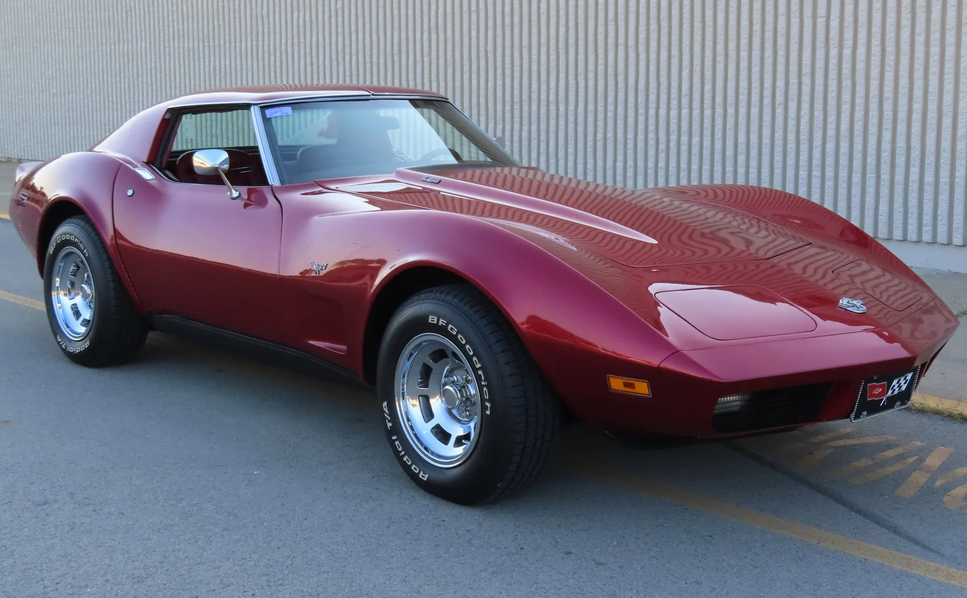 A dark red Corvette coupe with polished aluminum wheels and BFGoodrich white-letter tires, parked beside a ribbed wall and seen from the front right, with crossed-flags emblems on the nose and front fender.