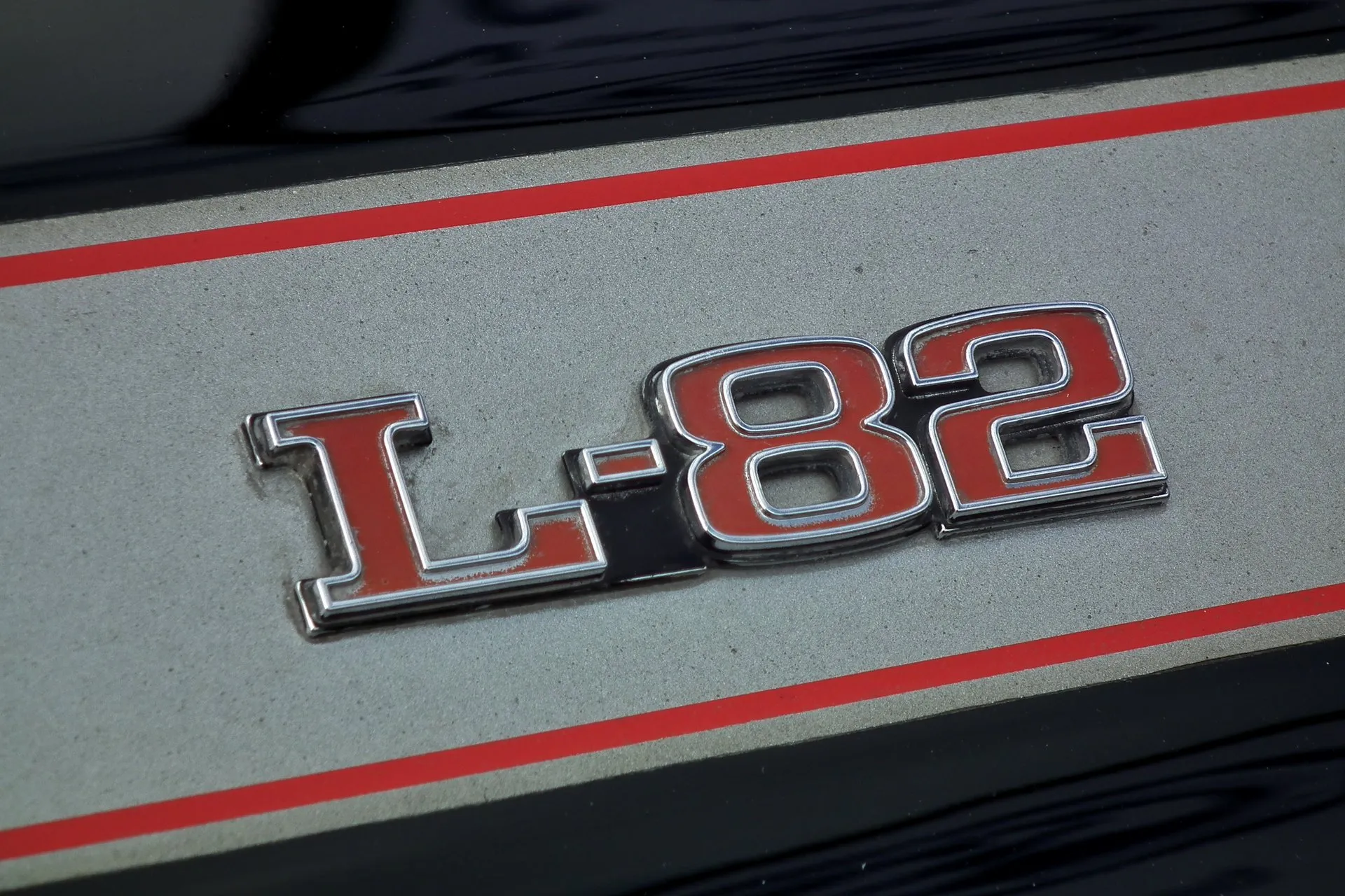 Close-up of a chrome L-82 emblem with red-filled letters on a silver hood stripe edged with red pinstripes, with black paint on either side.