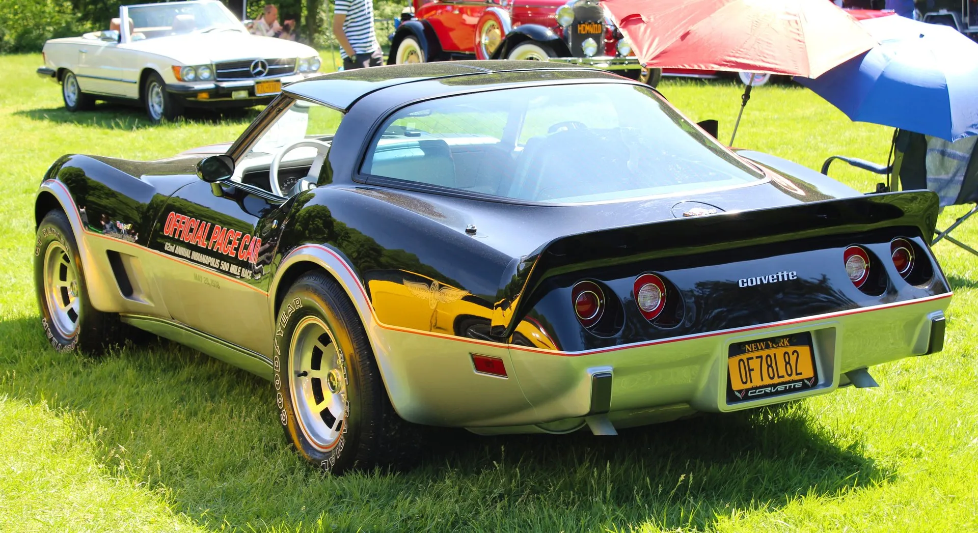The rear of a black-over-silver Corvette coupe with red pinstripes, a rear spoiler, a large fastback rear window, Official Pace Car lettering on the door and a winged-wheel emblem on the rear fender, parked on grass at a car show, with a New York plate reading OF78L82.