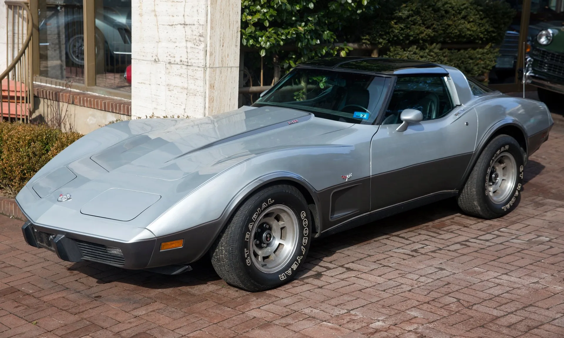 A two-tone silver Corvette coupe with a darker lower body and a pinstripe between the tones, an L82 emblem on the hood, aluminum wheels and white-letter Goodyear tires, parked on brick paving and seen from the front left.