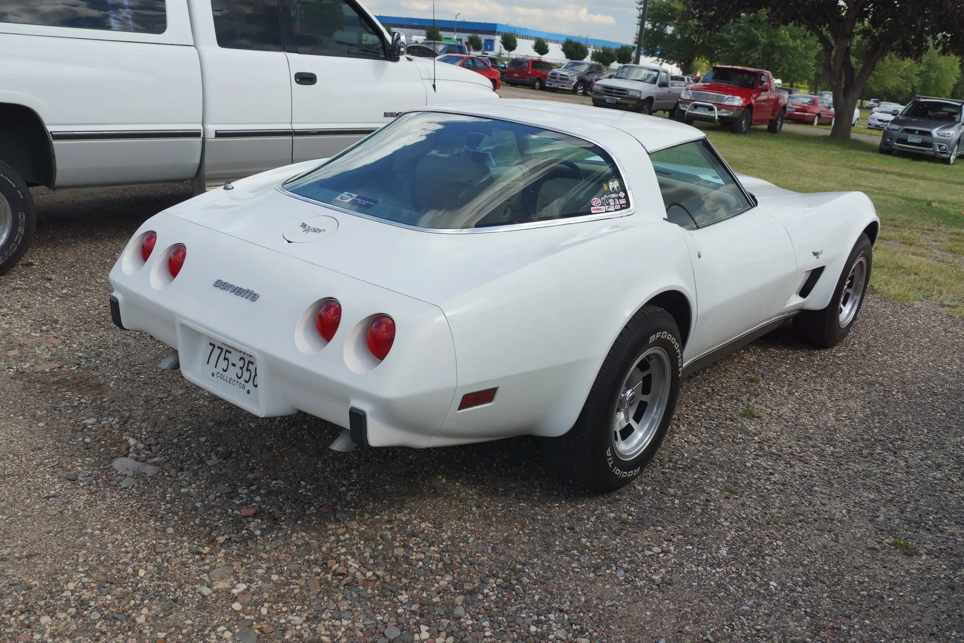 The rear of the same white Corvette coupe from the right, with a large fastback rear window, four round red taillights, a corvette script on the rear bumper cover, a crossed-flags emblem on the round fuel filler door and no rear spoiler.