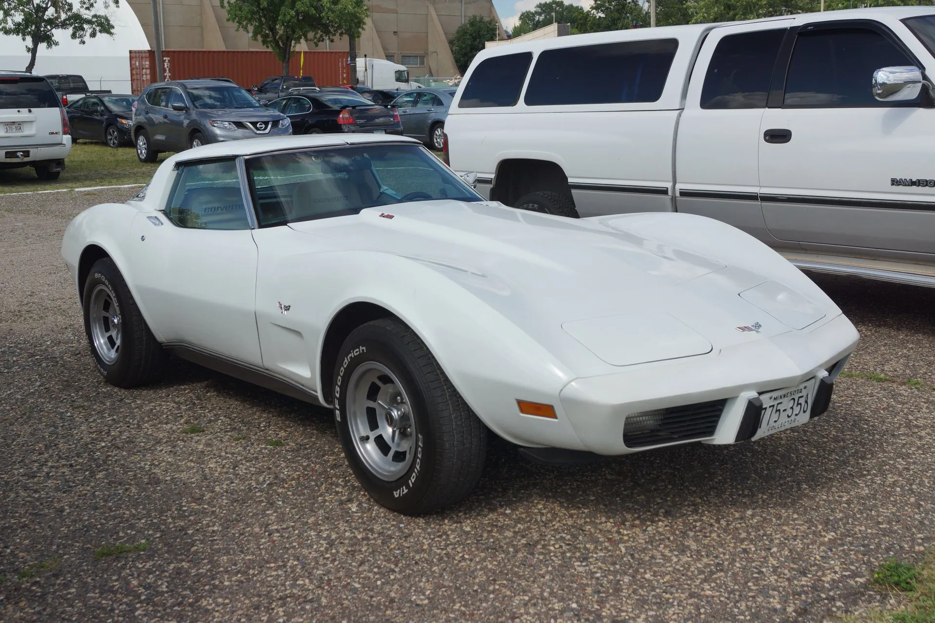 A white Corvette coupe with a fastback roof, seen from the front right on a gravel lot, with crossed-flags emblems on the nose and front fender, an L82 emblem at the back of the hood, slotted aluminum wheels and BFGoodrich white-letter tires, parked beside a white pickup.