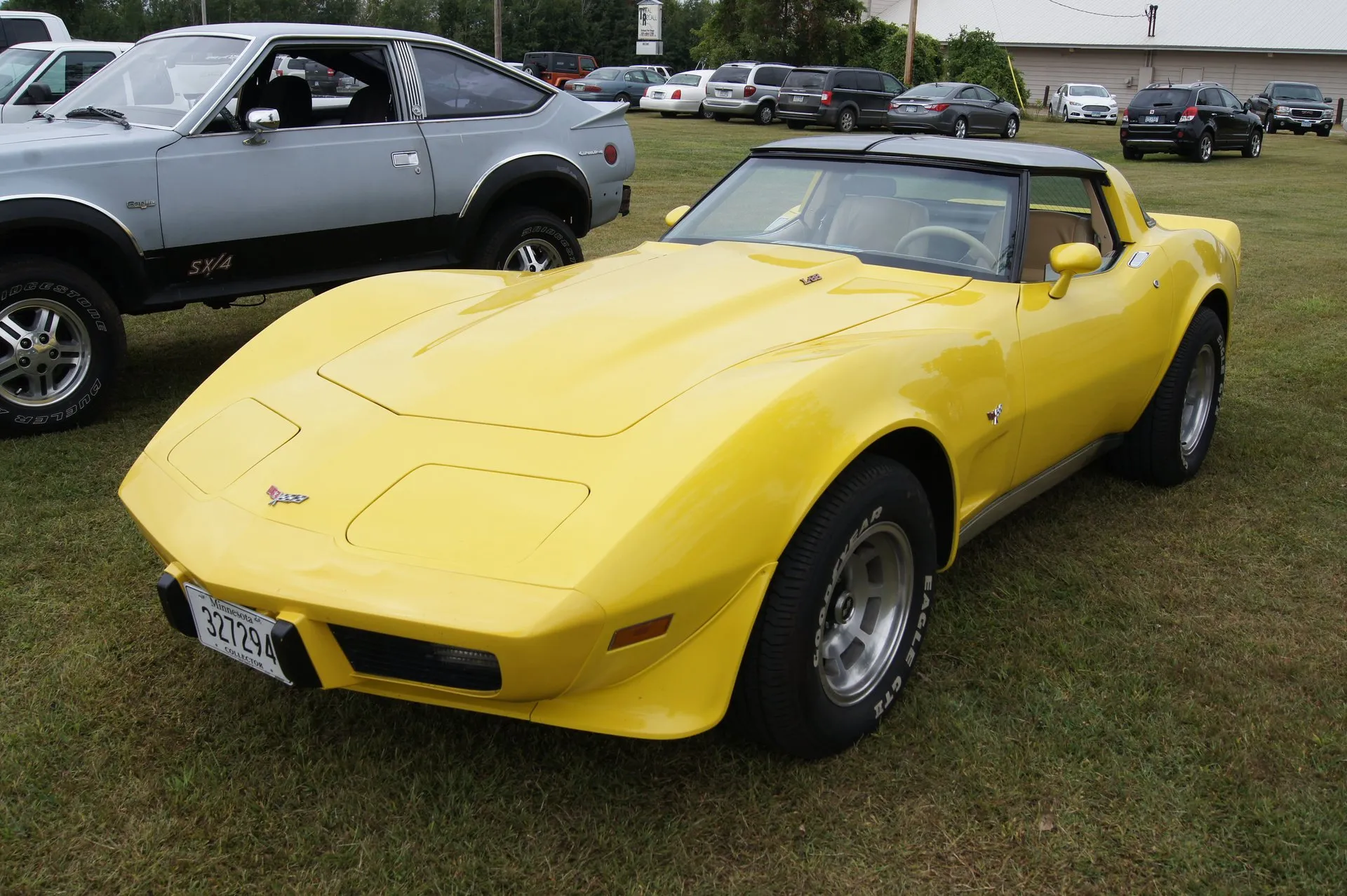 A yellow Corvette coupe with tinted roof panels and a beige interior, seen from the front left, parked on grass beside a silver AMC Eagle SX/4, with crossed-flags emblems on the nose and fender, an L82 emblem on the hood, slotted aluminum wheels and Goodyear white-letter tires.