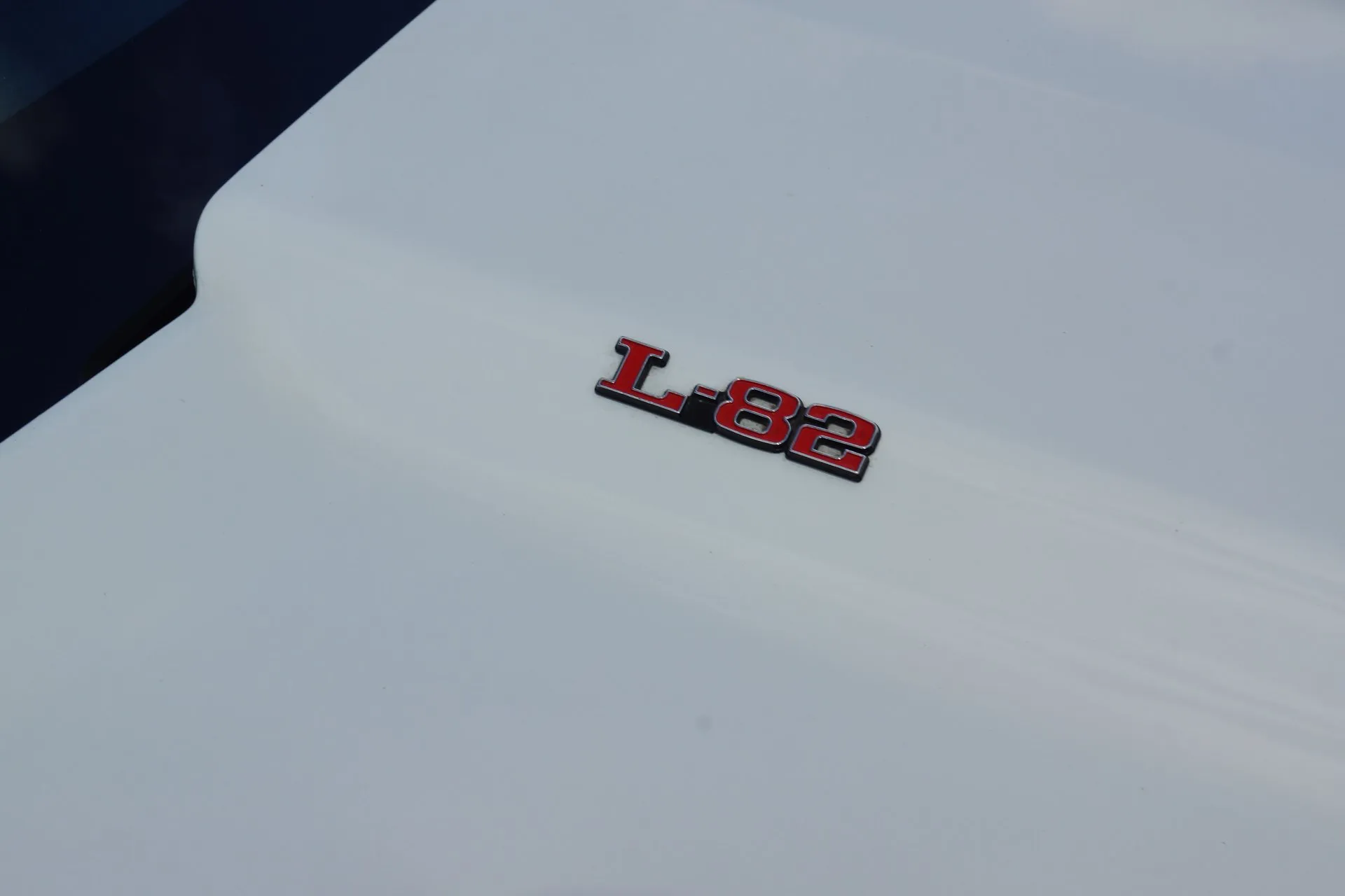 Close-up of a chrome L-82 emblem with red-filled letters on a white Corvette hood, near the base of the windshield.