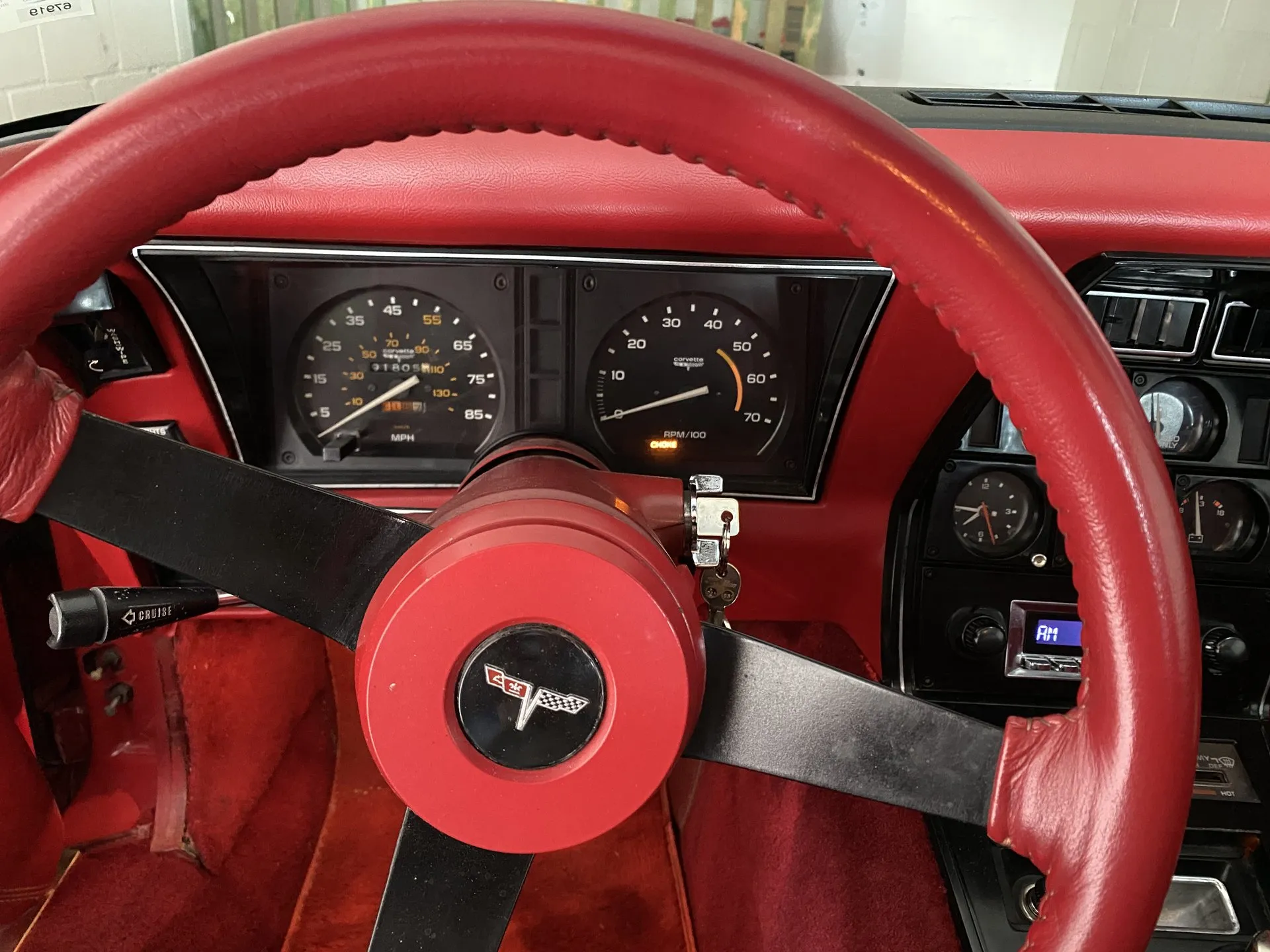 The instruments of a C3 Corvette with red trim, seen past a red leather-wrapped steering wheel: a speedometer marked to 85 mph with 55 picked out and an inner kilometer scale, beside a tachometer marked to 7,000 rpm with its CHOKE light lit.