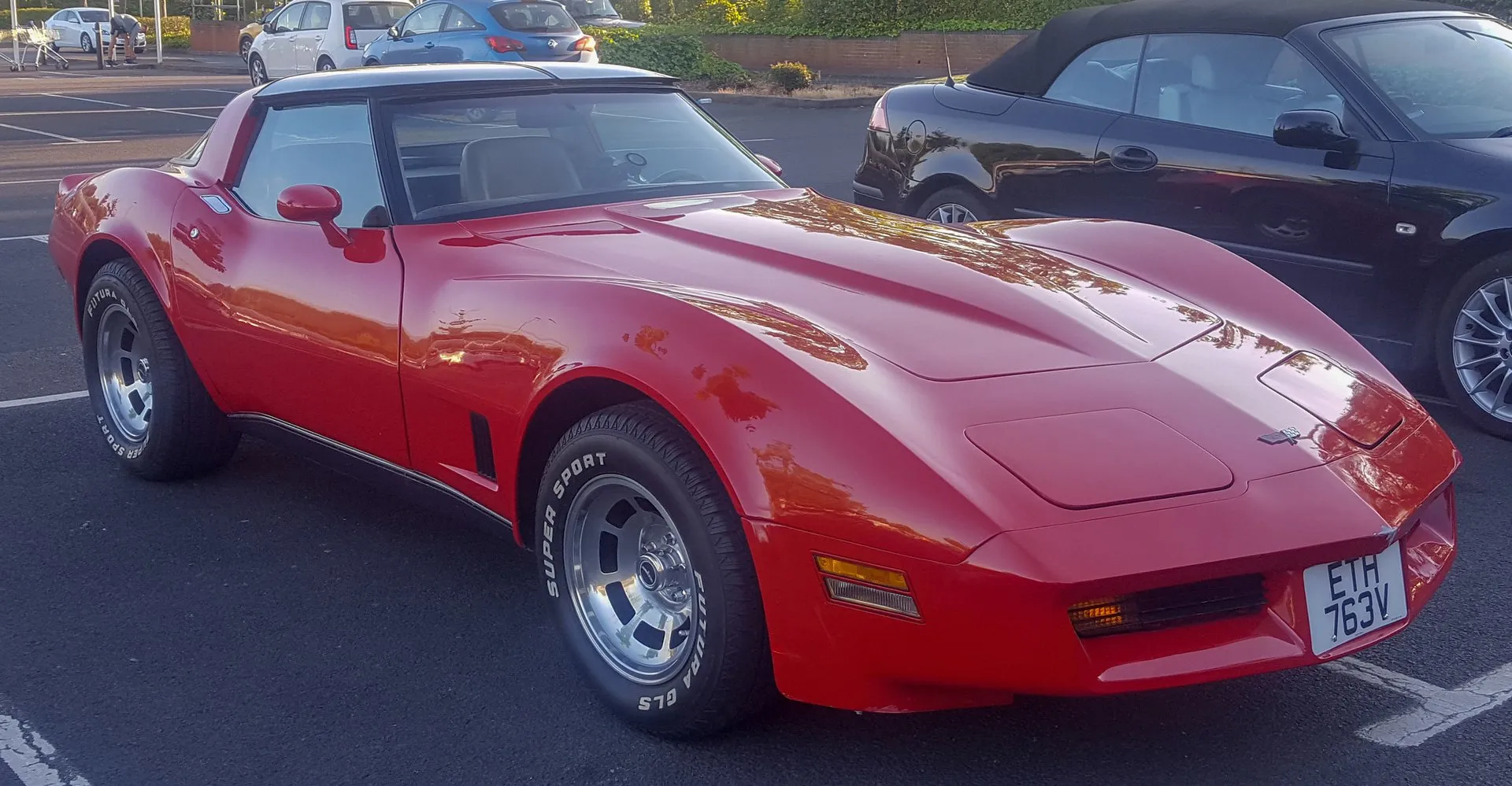 A red Corvette coupe with tan seats in a parking lot, seen from the front and side, with a spoiler molded into the front bumper cover, a recessed grille, black louvers behind the front wheel, aluminum wheels and a British number plate.