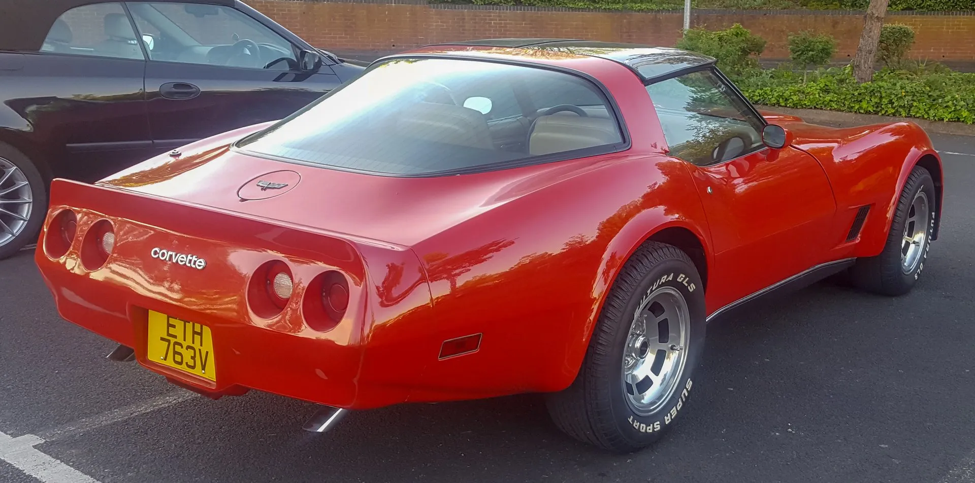 The rear of the same red Corvette coupe, seen from behind and to the right, with a spoiler molded into the rear bumper cover, a Corvette nameplate between four round taillights, crossed flags on the fuel door, twin exhaust tips and a yellow British number plate.