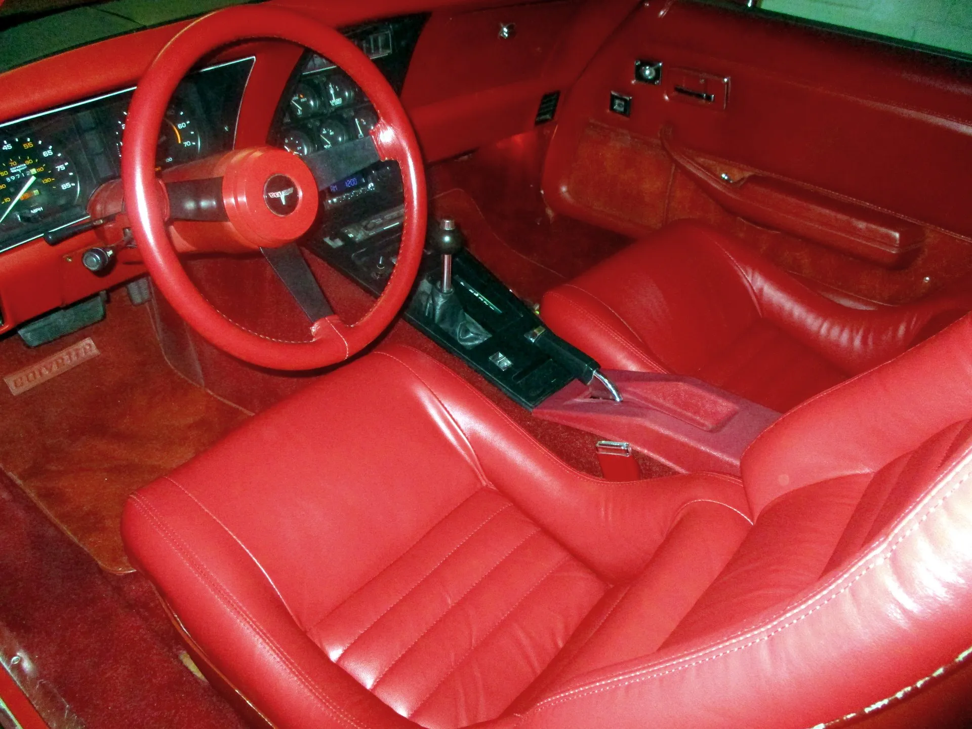 The cabin of a C3 Corvette with red leather seats, red carpet and door panels, a red leather-wrapped steering wheel and the automatic transmission's shifter on the center console.