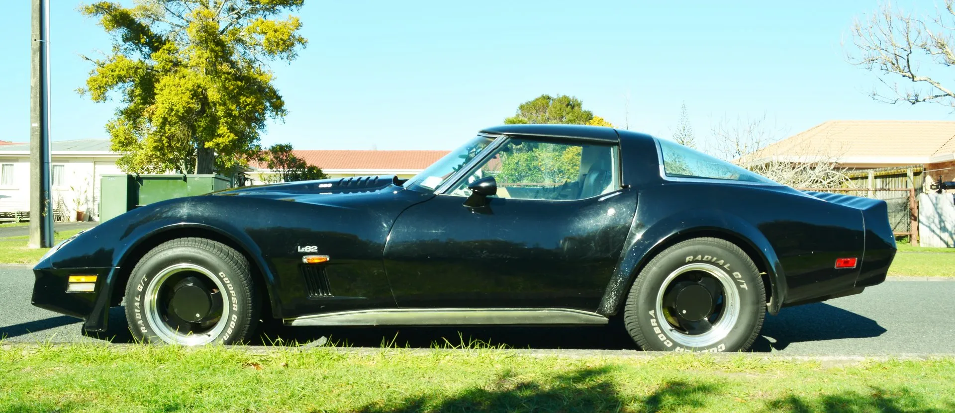 A black Corvette coupe in side profile on a suburban street, with an L82 emblem on the front fender above the side marker light, black louvers behind the front wheel, louvers cut into the hood and aftermarket wheels.