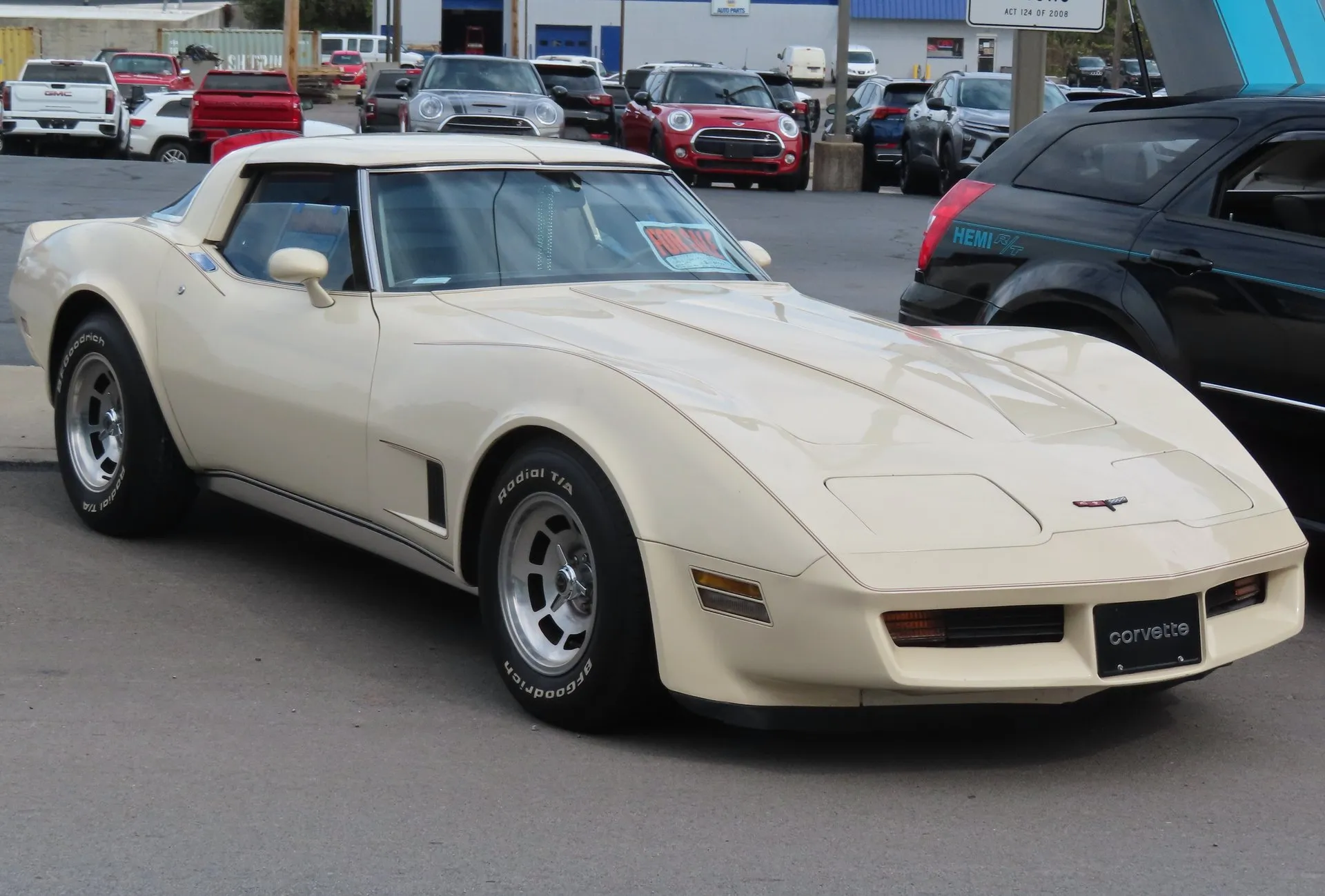 A beige Corvette coupe seen from the front right in a parking lot, with a for-sale sign inside the windshield, aluminum wheels and BFGoodrich white-letter tires.