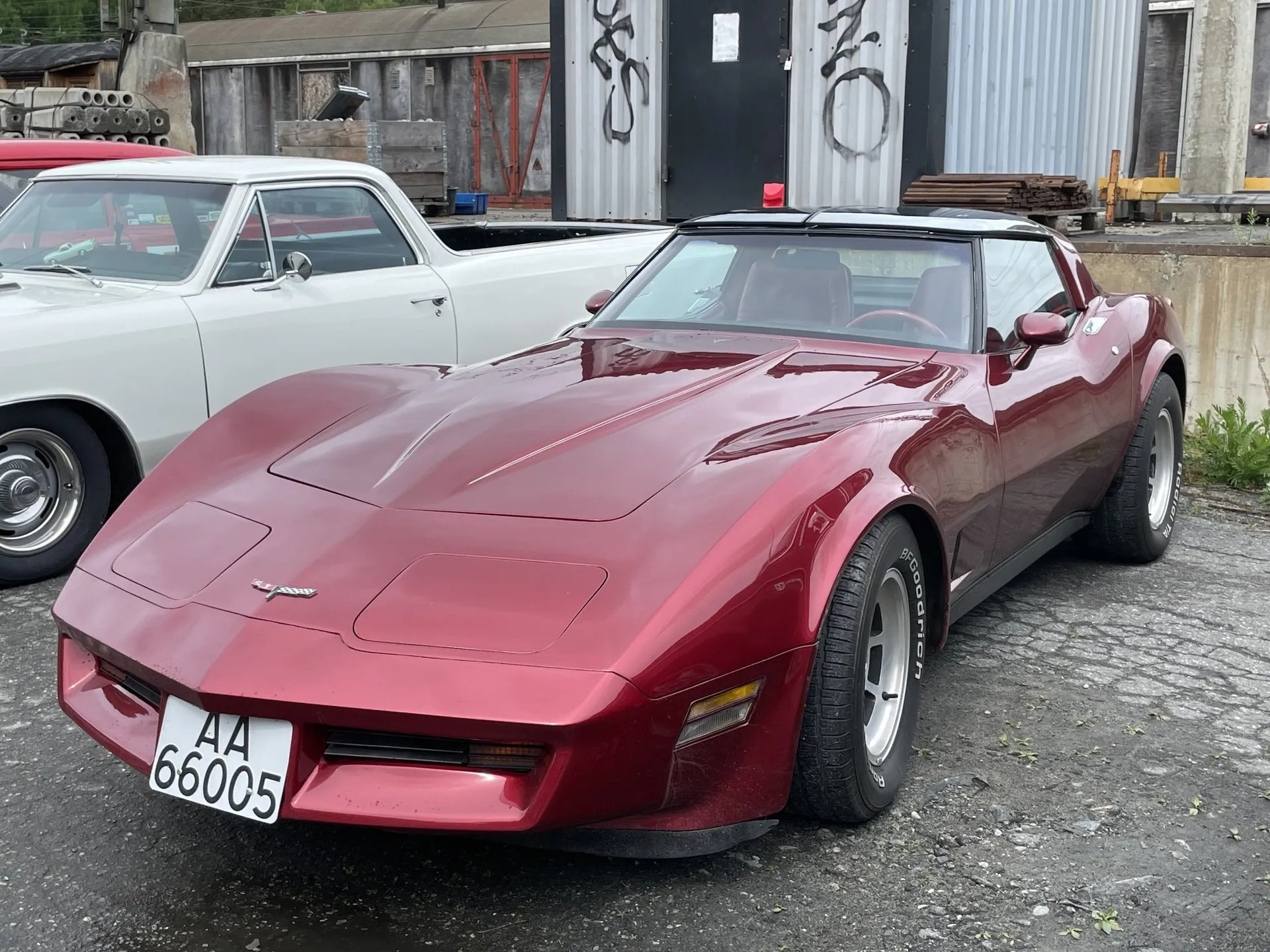 A dark red metallic Corvette coupe with black roof panels parked on broken pavement beside a white classic car, seen from the front left, with a white Norwegian number plate and BFGoodrich tires.