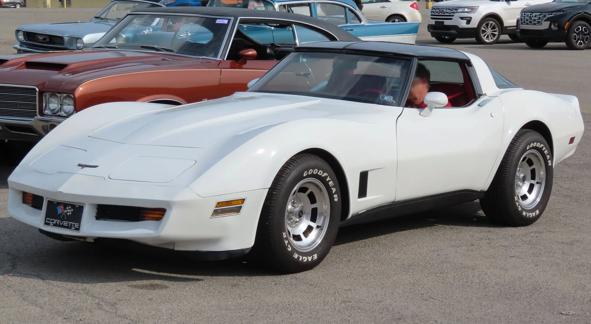 A white Corvette coupe with black roof panels and red seats at an outdoor car show, seen from the front left, with a spoiler molded into the nose, black louvers behind the front wheel, aluminum wheels and Goodyear white-letter tires.