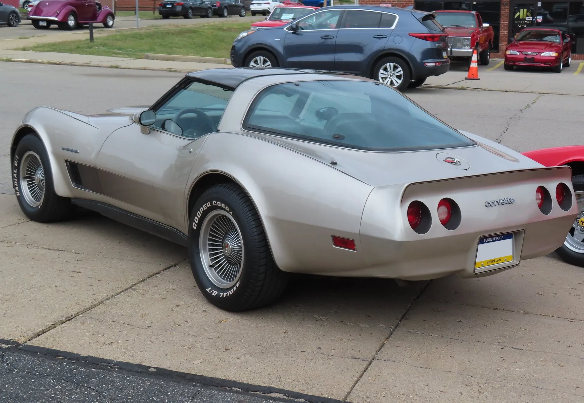 A silver-beige Corvette coupe in a parking lot, seen from the rear left, showing its large rear glass, darker shaded side graphics, finned multi-spoke wheels on white-letter tires and a Corvette nameplate between four round taillights.