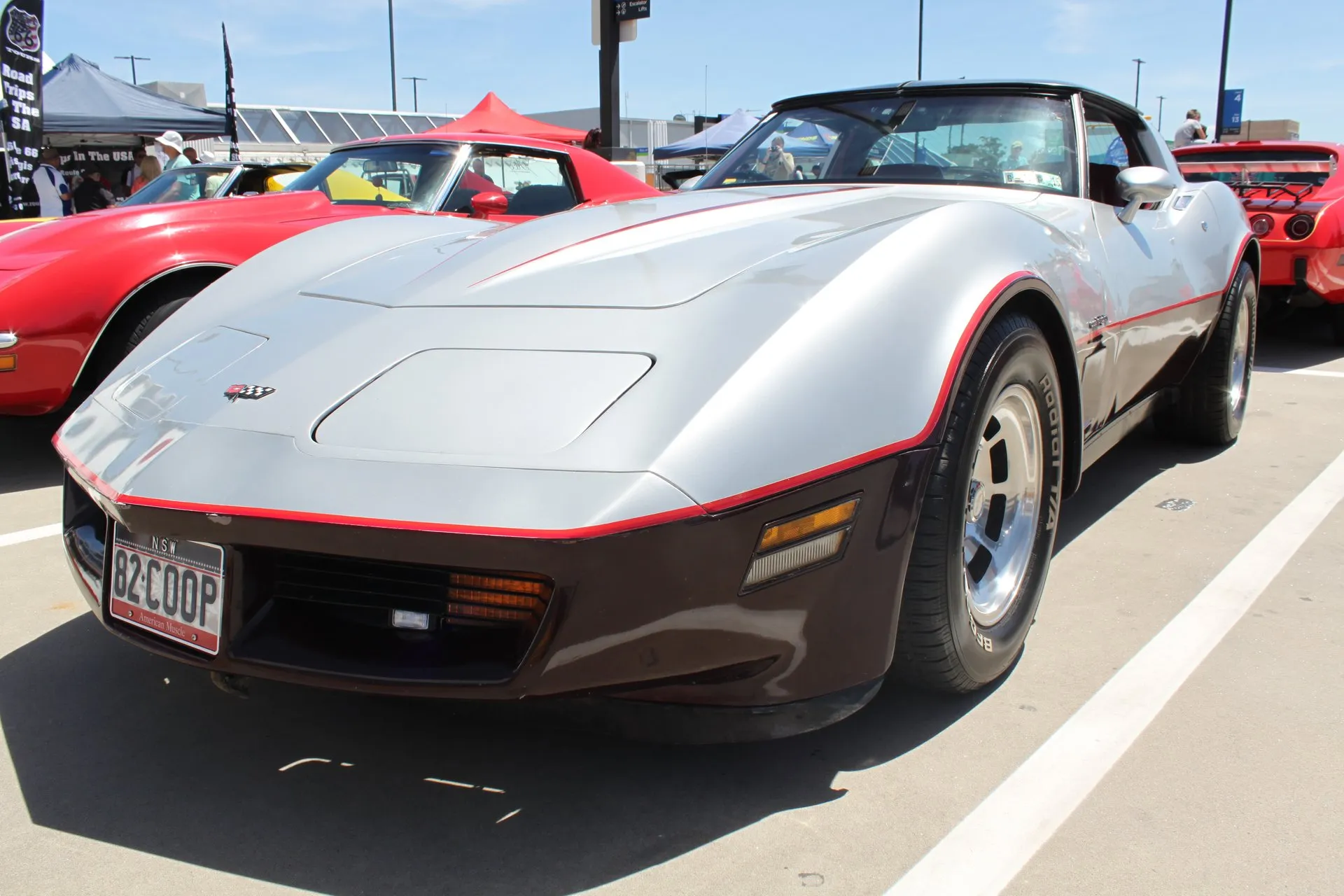 A silver and dark red two-tone Corvette coupe with a red pinstripe along the color break, seen from the front left at an outdoor car show, with aluminum wheels, white-letter tires and a New South Wales number plate reading 82 COOP.