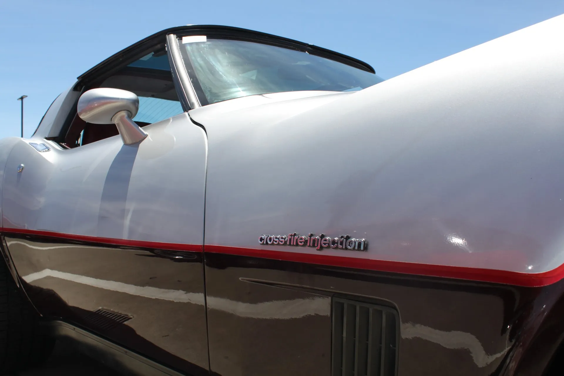 Close-up of a silver and dark red Corvette's front fender and door, with a chrome script reading cross-fire injection on the fender above a red pinstripe and a black louvered vent.