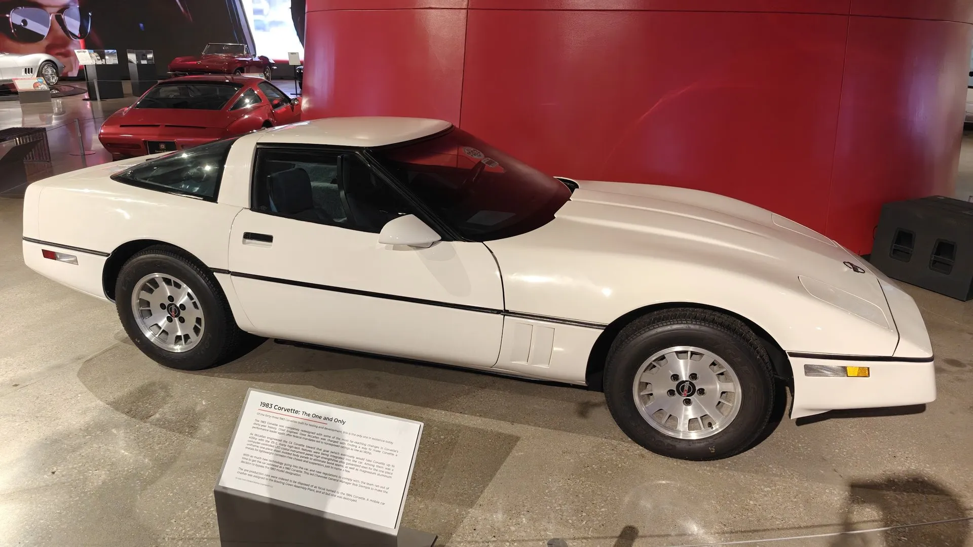 A white Corvette coupe of the C4 shape in side view on a museum floor, in front of a curved red wall, with slotted aluminum wheels and a placard beside it titled 1983 Corvette: The One and Only.
