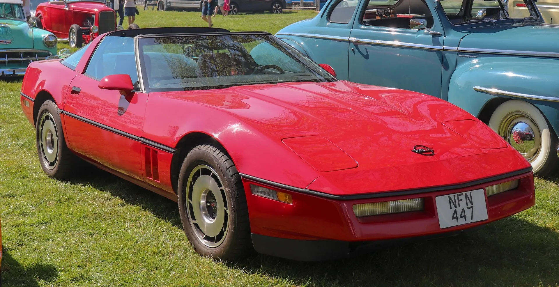 A red C4 Corvette coupe with a dark roof panel and turbine-style aluminum wheels, parked on grass at a car show beside an older blue sedan, seen from the front right, with a British number plate.