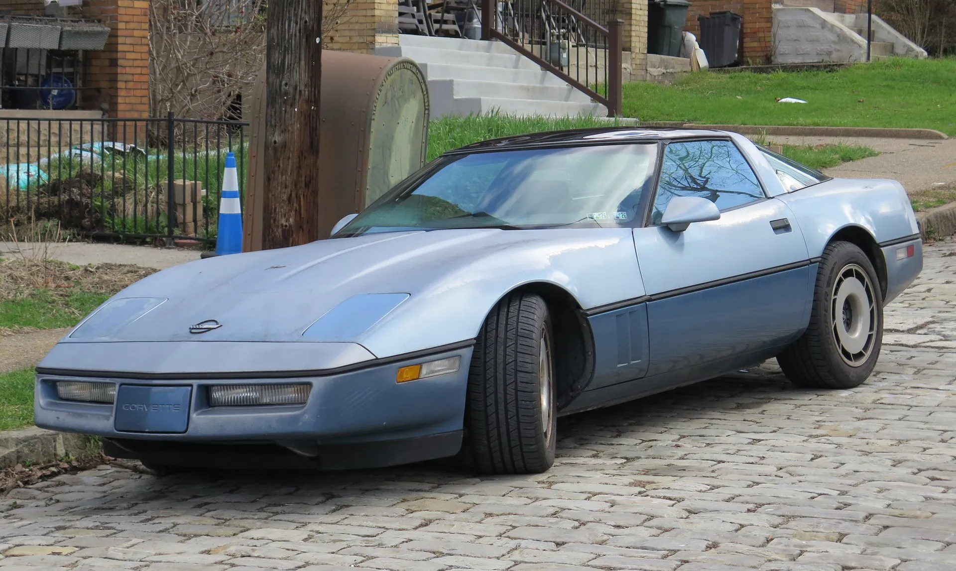 A Corvette coupe in light blue over a darker blue lower body, parked on a cobblestone street in front of brick houses, seen from the front left, with its headlamps closed, a dark roof panel and turbine-style aluminum wheels. The paint is dusty and weathered.