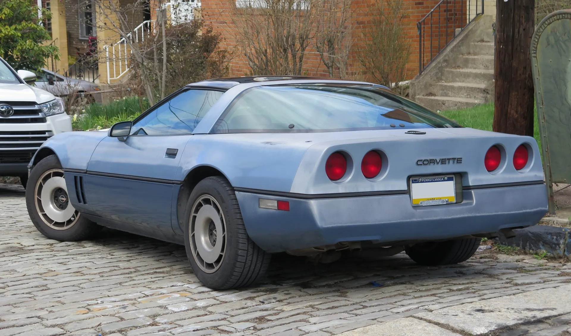 The rear of the light blue two-tone Corvette coupe on the cobblestone street, seen from the rear left, showing the large glass hatch, four round red taillamps, Corvette lettering on the rear panel and a Pennsylvania plate.