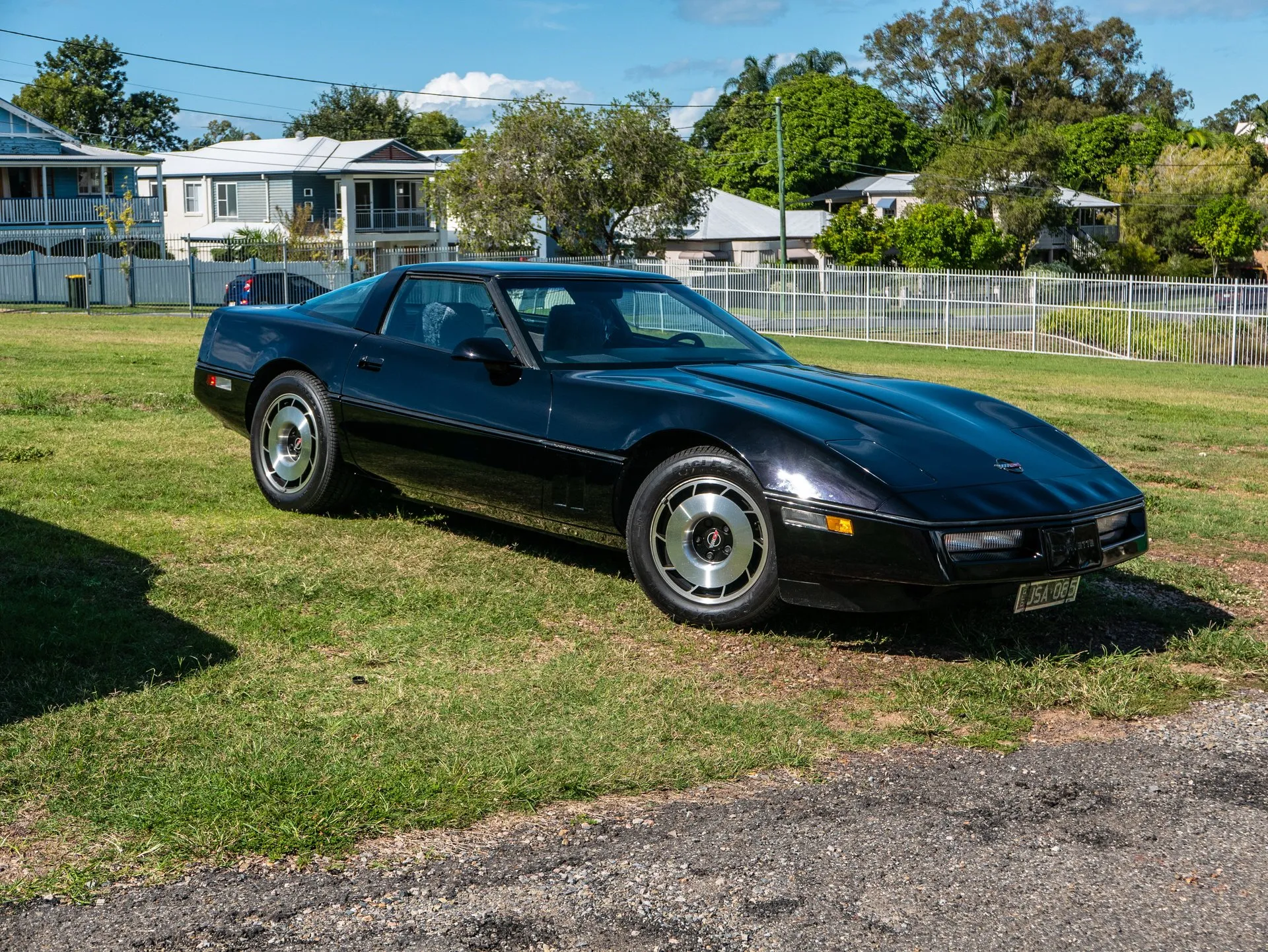 Black 1985 Chevrolet Corvette coupe parked on grass in front of a white fence and houses, seen from the front right, with a script on the side molding behind the front wheel.