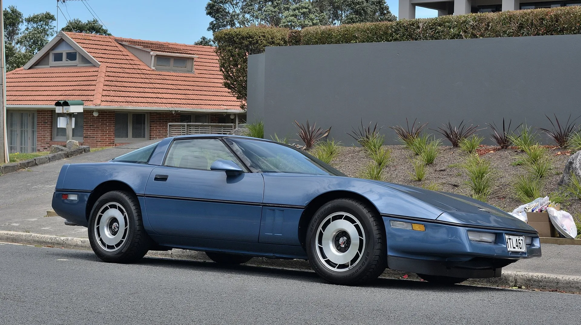Blue 1985 Chevrolet Corvette coupe parked at a curb on a suburban street, seen from the front right, with Tuned Port Injection script on the side molding and BFGoodrich tires.