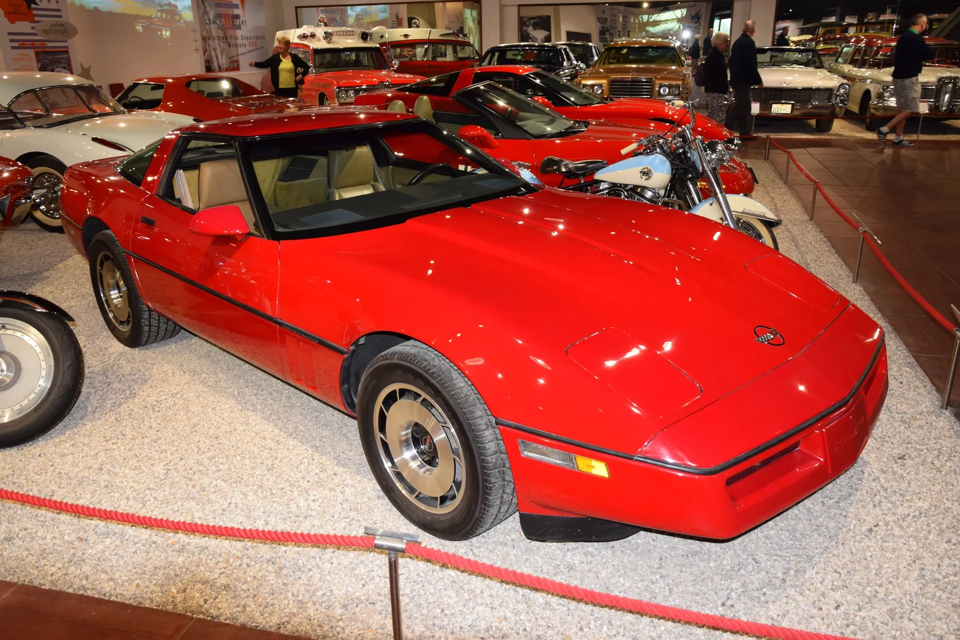 Bright red 1985 Chevrolet Corvette coupe with a saddle interior, on display behind a rope in a car museum, seen from the front right.