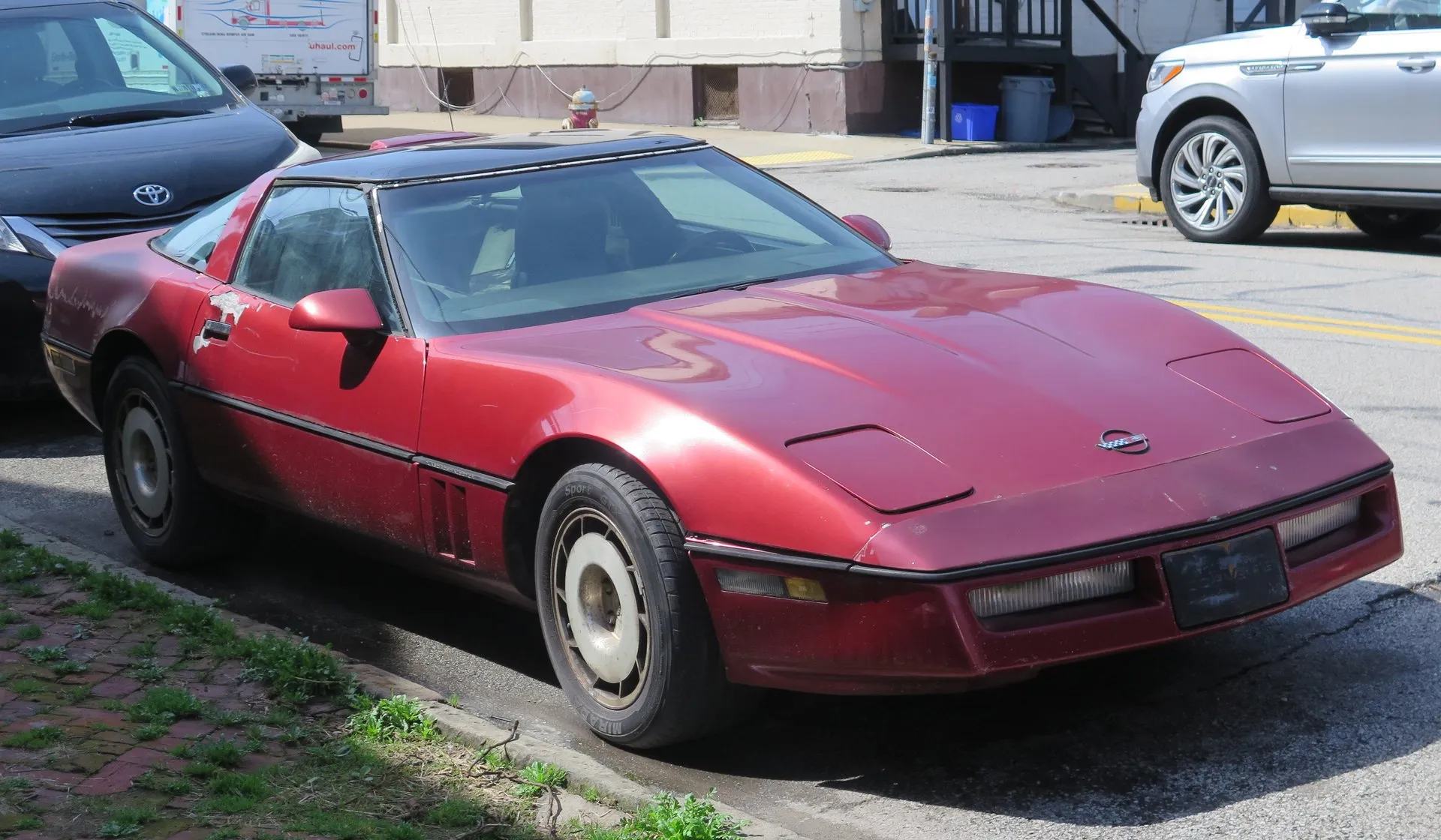 The same dark red 1986 Chevrolet Corvette coupe seen from the front right, headlamps closed, with faded and peeling paint on the hood and door.