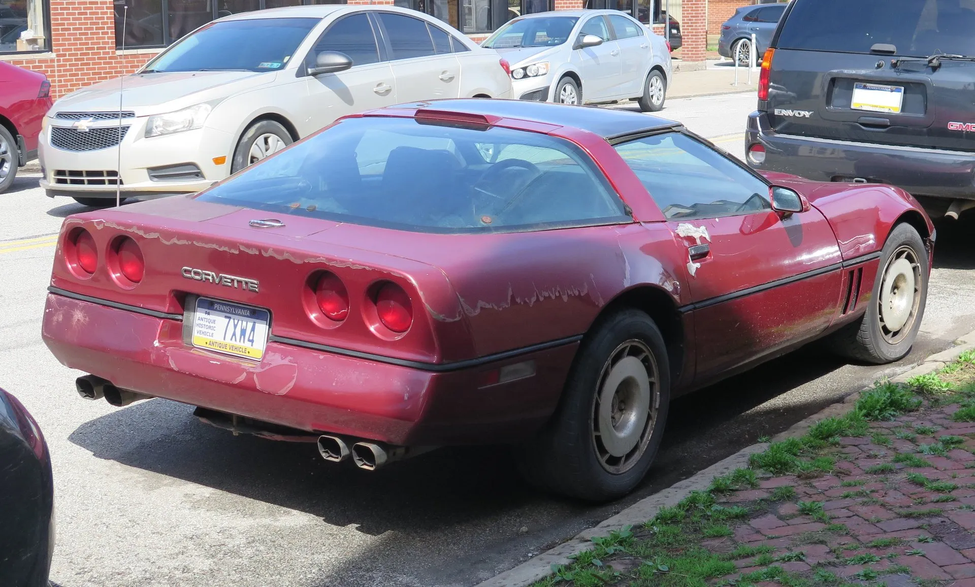 Dark red 1986 Chevrolet Corvette coupe parked at a curb, seen from the rear right, with a third brake light at the top of the rear glass, four round taillights, quad exhaust tips and clearcoat peeling from the hatch, bumper and door.