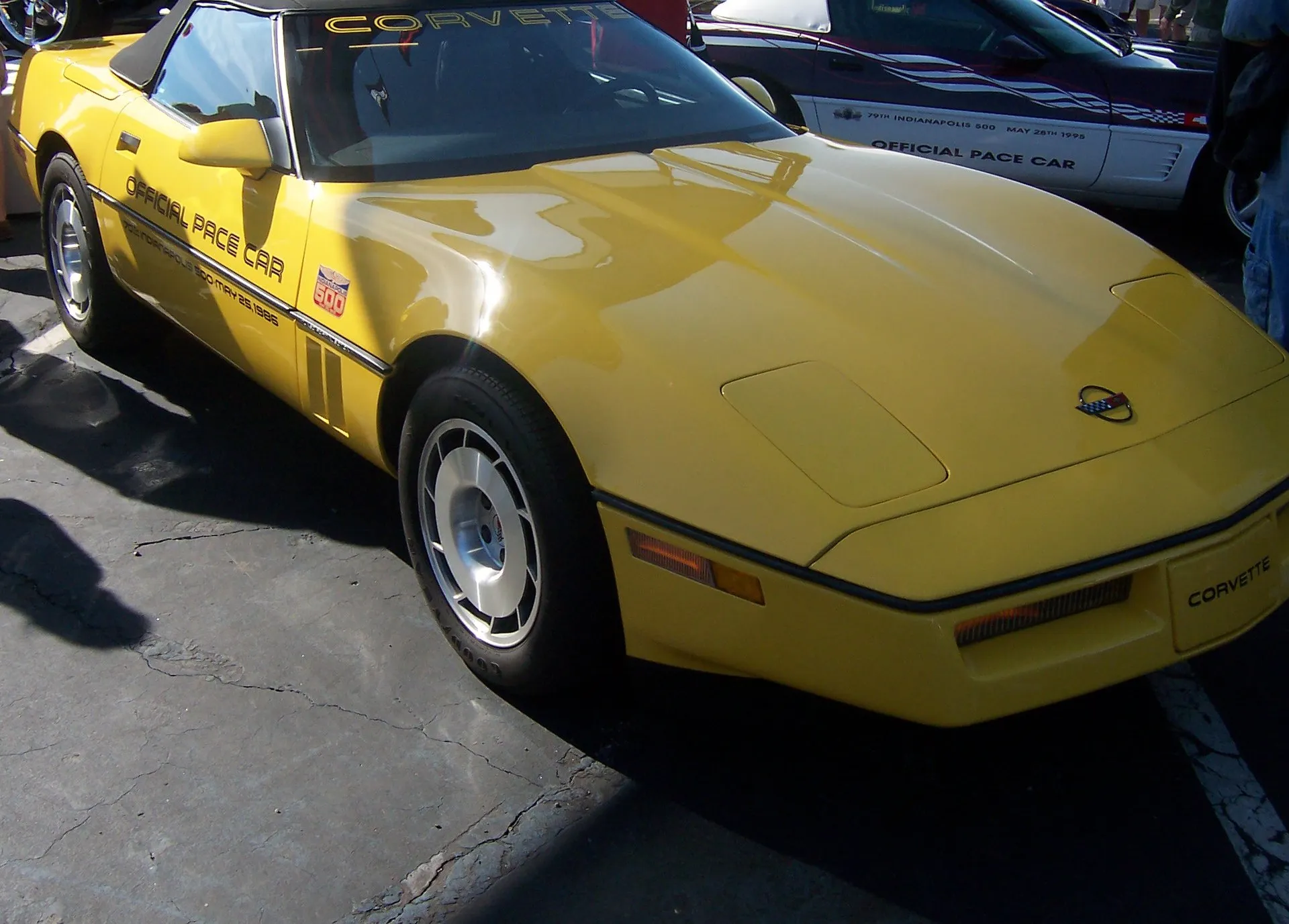 Yellow 1986 Chevrolet Corvette convertible with its black top up, Official Pace Car lettering and an Indianapolis 500 logo on the door, and silver factory wheels, parked in a lot beside a white and purple 1995 pace car.