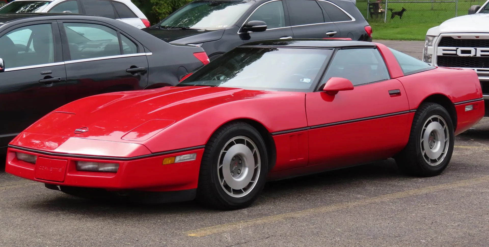 Bright red 1987 Chevrolet Corvette coupe with a dark roof panel, parked on asphalt among modern cars, seen from the front left, with silver factory wheels and gray wheel centers.
