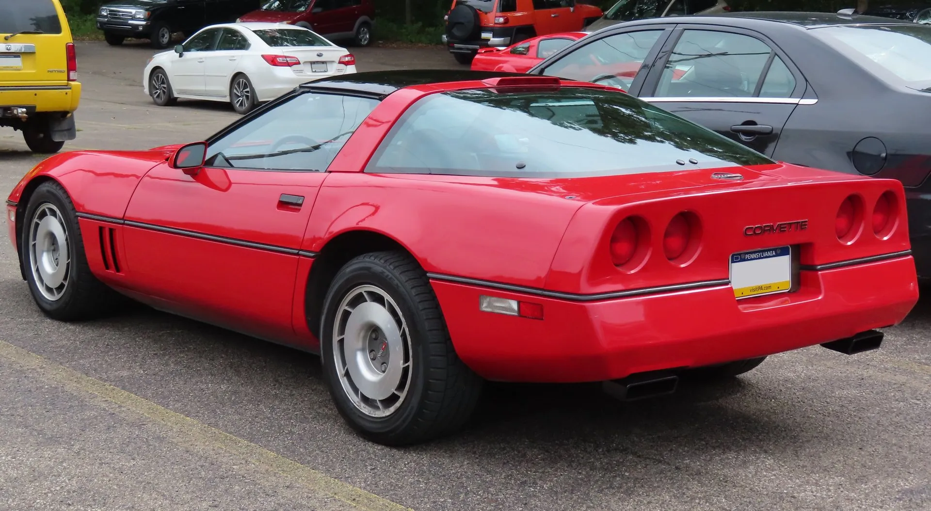 The same red 1987 Corvette coupe seen from the rear left, with four round taillights, a brake light at the top of the rear glass and twin exhaust outlets under the bumper.