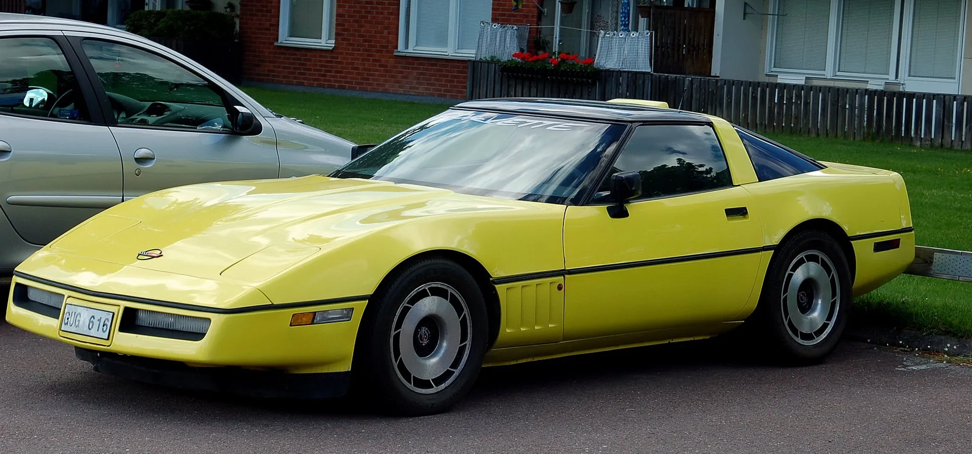 Yellow C4 Corvette coupe with a dark tinted roof panel and silver wheels, parked at a curb on a Swedish residential street beside a gray hatchback.