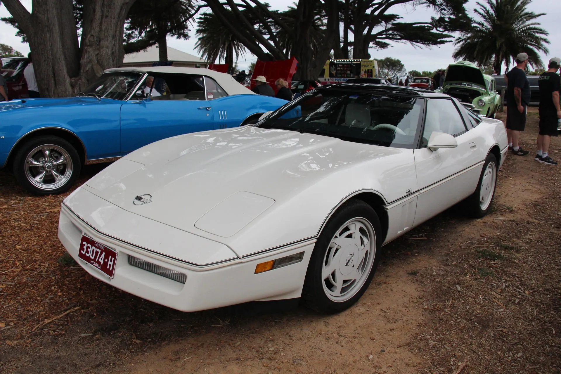 White 1988 Corvette 35th Anniversary coupe with white-painted slotted wheels, white side moldings and a dark roof, parked on a dirt lot at a car show beside a blue convertible, seen from the front left.