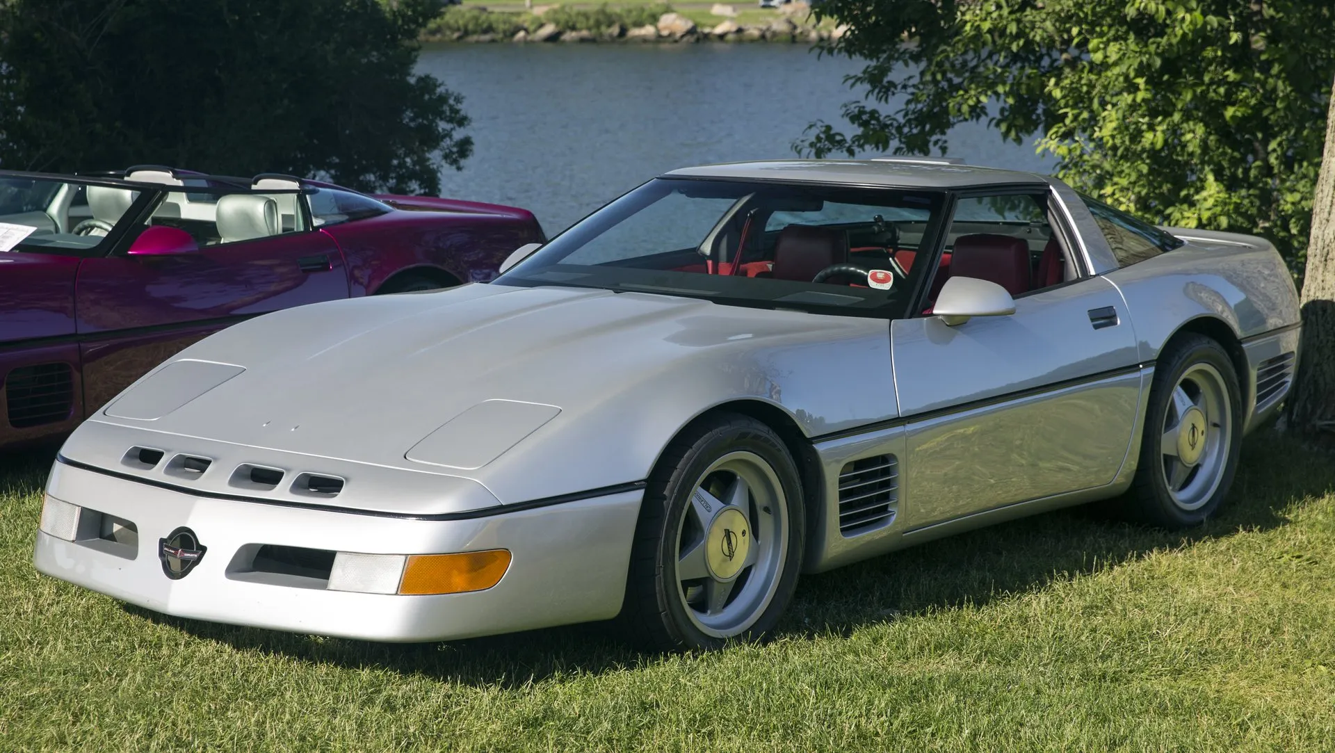 Silver Corvette coupe with a Callaway badge on a slotted nose, slatted side vents, five-spoke wheels and a red interior, parked on grass by a lake, seen from the front left.