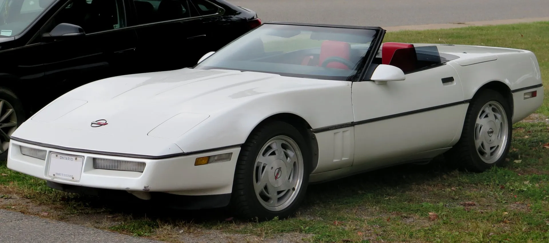 White 1988 Corvette convertible with its top down and a red interior, parked on grass beside a road, seen from the front left, on silver wheels with six slanted vents.