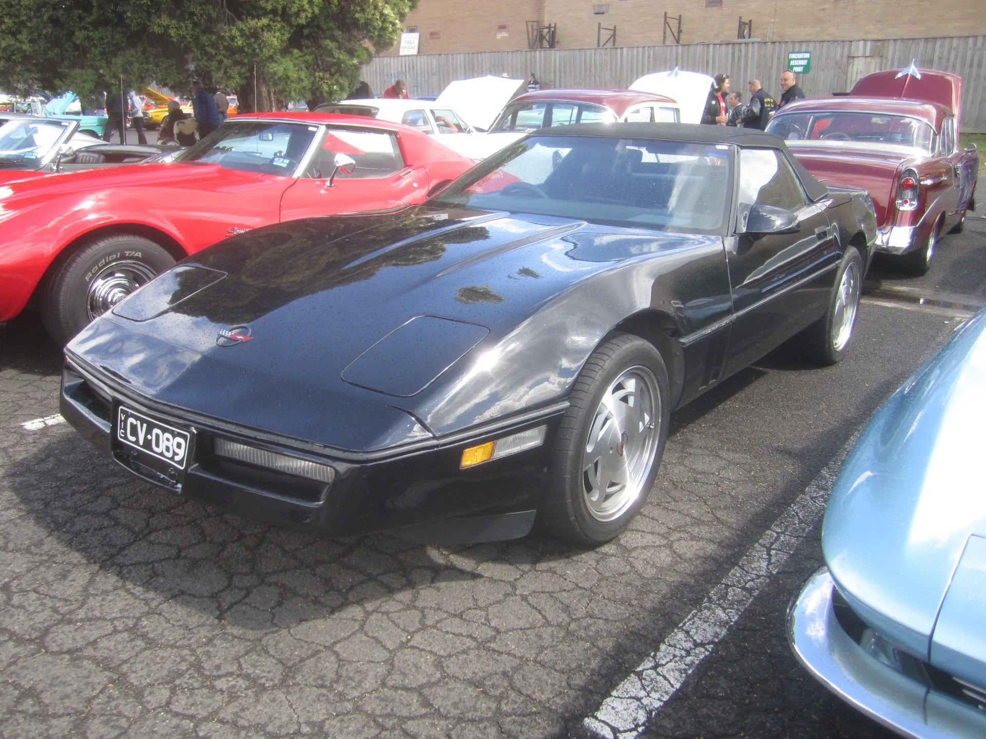 Black C4 Corvette convertible with its black soft top up and silver slotted wheels, parked at an outdoor car show among older cars, seen from the front left.