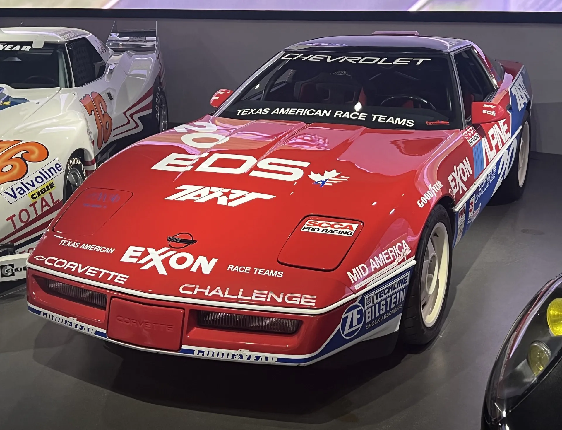 Red Corvette Challenge race car numbered 20, with EDS and Exxon lettering on the hood, Corvette Challenge on the nose and blue and white sponsor decals on the side, on display in a museum beside other race cars.