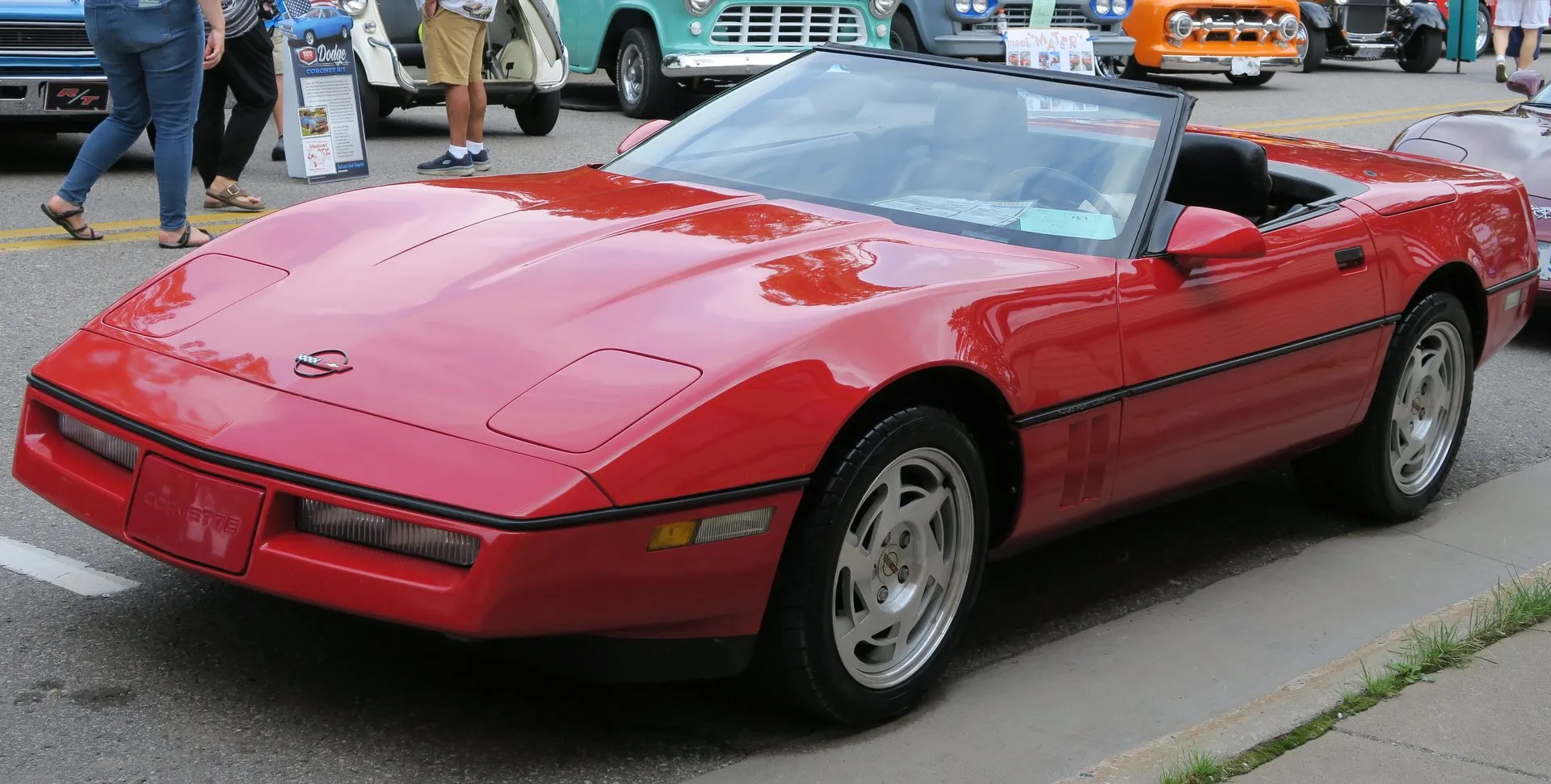 Bright red Chevrolet Corvette C4 convertible with the top down and a black interior, parked at a street car show and seen from the front left, with silver slotted wheels and onlookers' legs at the edge of the frame.