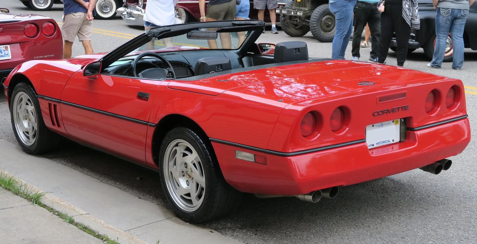 The same red convertible from the rear left with its top folded, showing four round taillamps on a concave rear panel, a small stop lamp at the top of the rear fascia, Corvette lettering, a historical license plate and four exhaust tips.