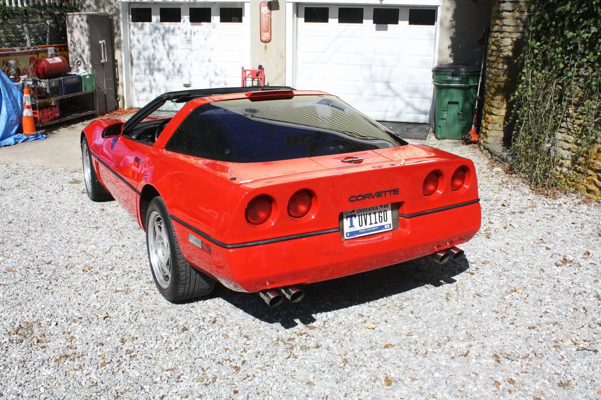 Red Chevrolet Corvette C4 coupe with its roof panel off, seen from the rear left on a gravel driveway, showing four round taillamps on a concave tail, a stop lamp at the top of the rear glass, Corvette lettering, an Indiana plate and four exhaust tips.