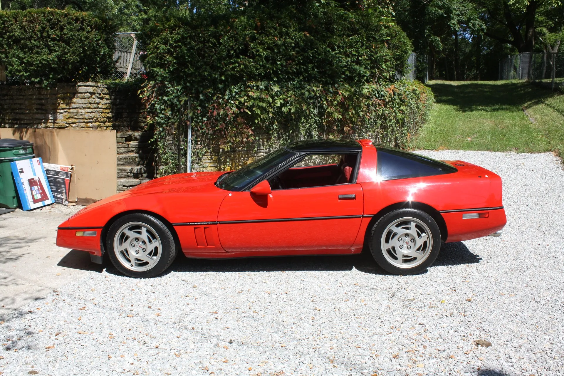 Red Chevrolet Corvette C4 coupe with a dark roof panel, seen from its left side on a gravel driveway, with a red interior, silver slotted wheels and Tuned Port Injection lettering on the side molding behind the front wheel.