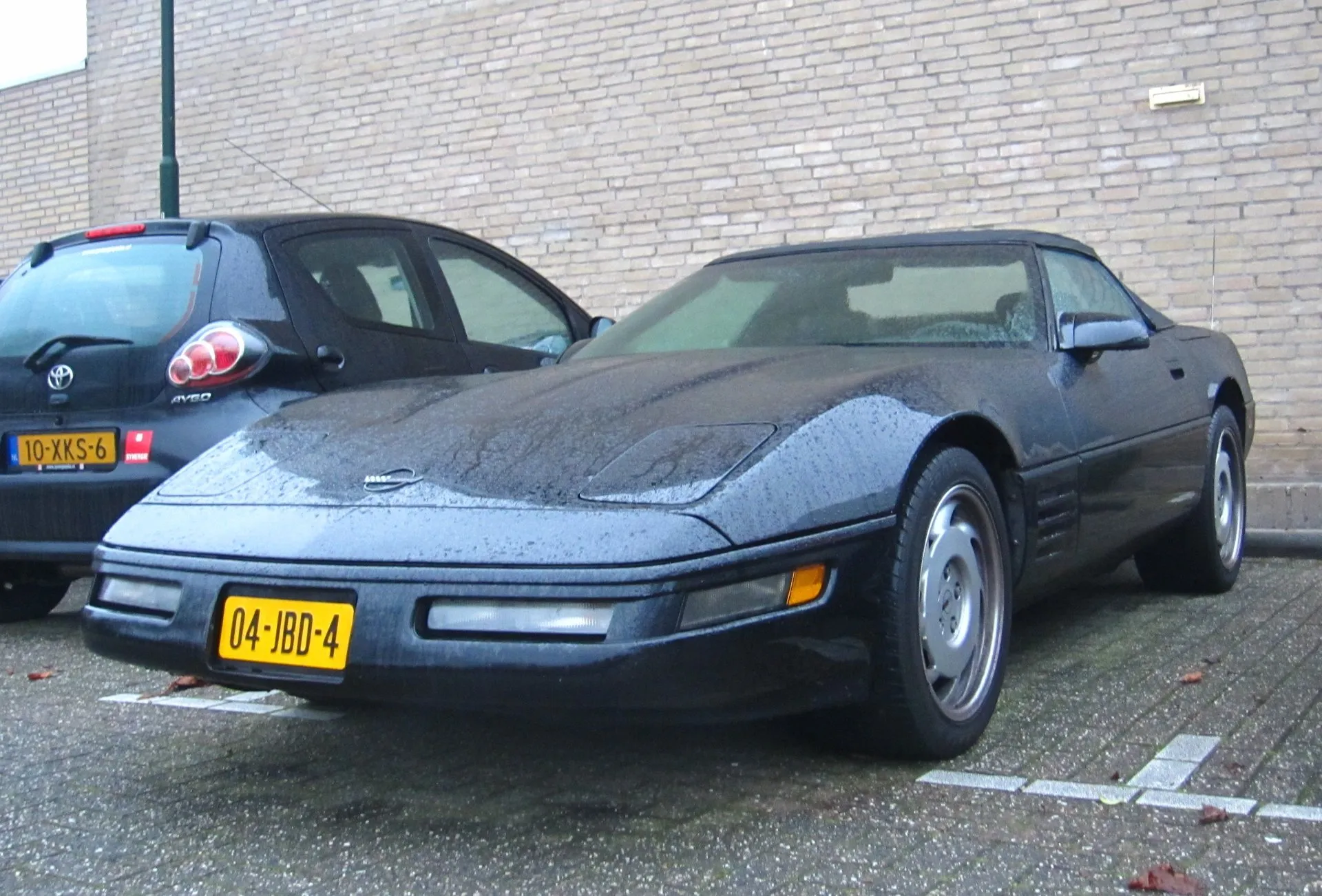 Dark-colored Chevrolet Corvette C4 convertible with its black top up, wet with rain, parked beside a small hatchback in front of a brick wall, with a Dutch registration plate.