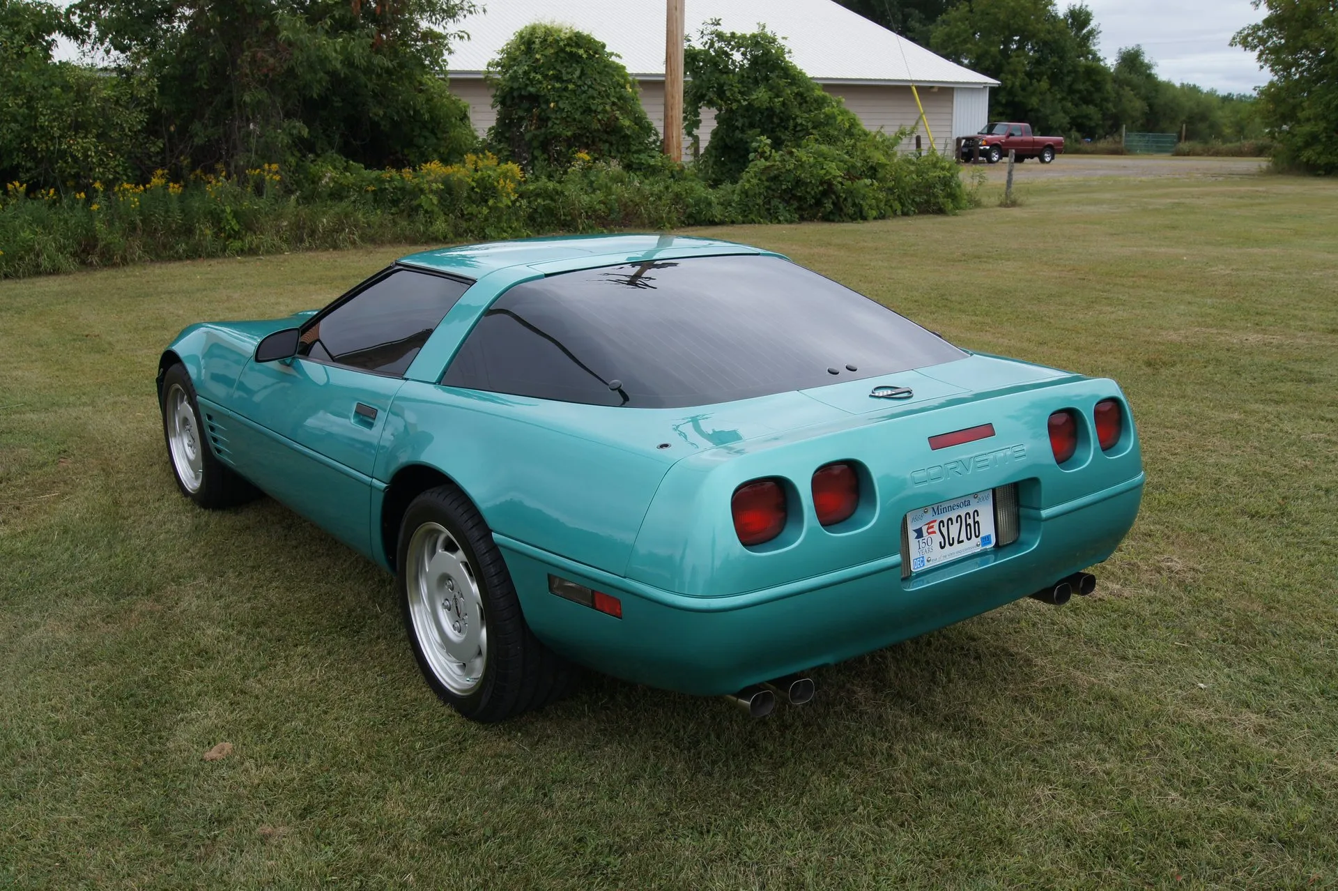 Turquoise Metallic Chevrolet Corvette C4 coupe parked on grass, seen from the rear left, with a convex rear bumper, four square red taillamps, a stop lamp above the license plate and four round exhaust tips.