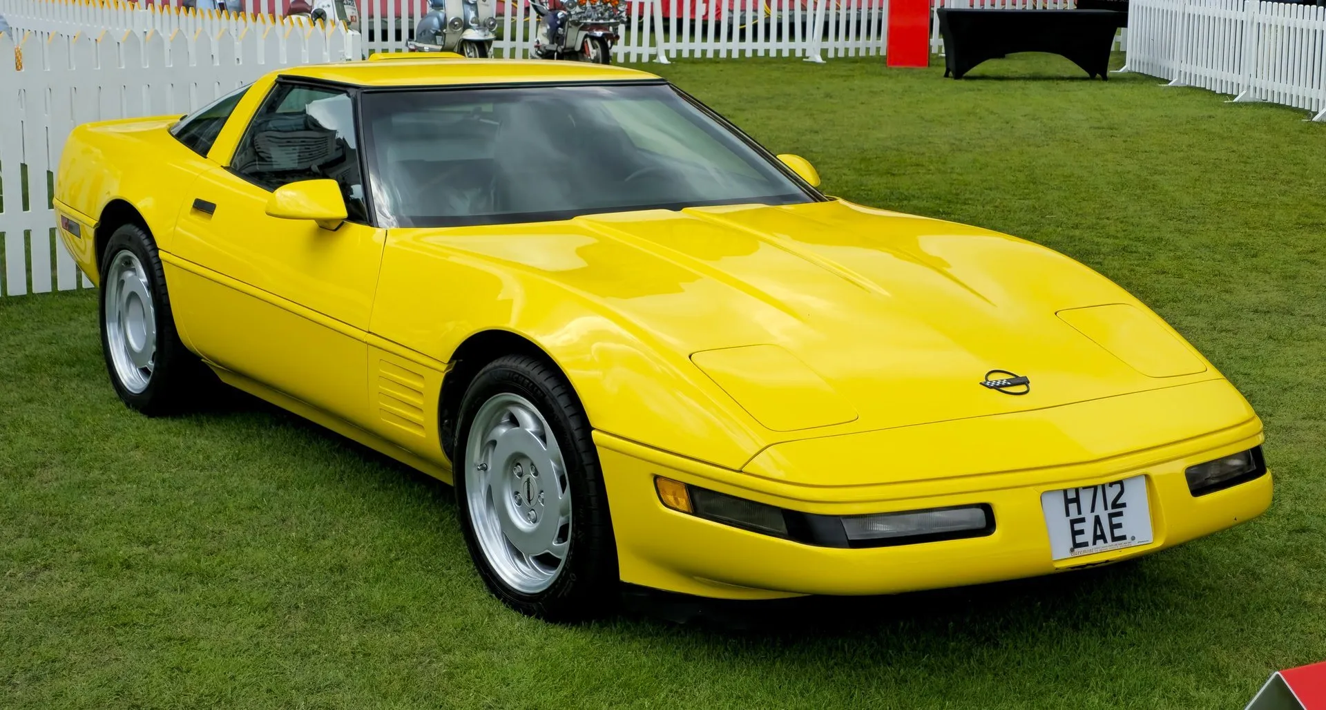 Yellow Chevrolet Corvette C4 coupe parked on grass at a show behind a white picket fence, seen from the front right, with silver slotted wheels and a British registration plate.