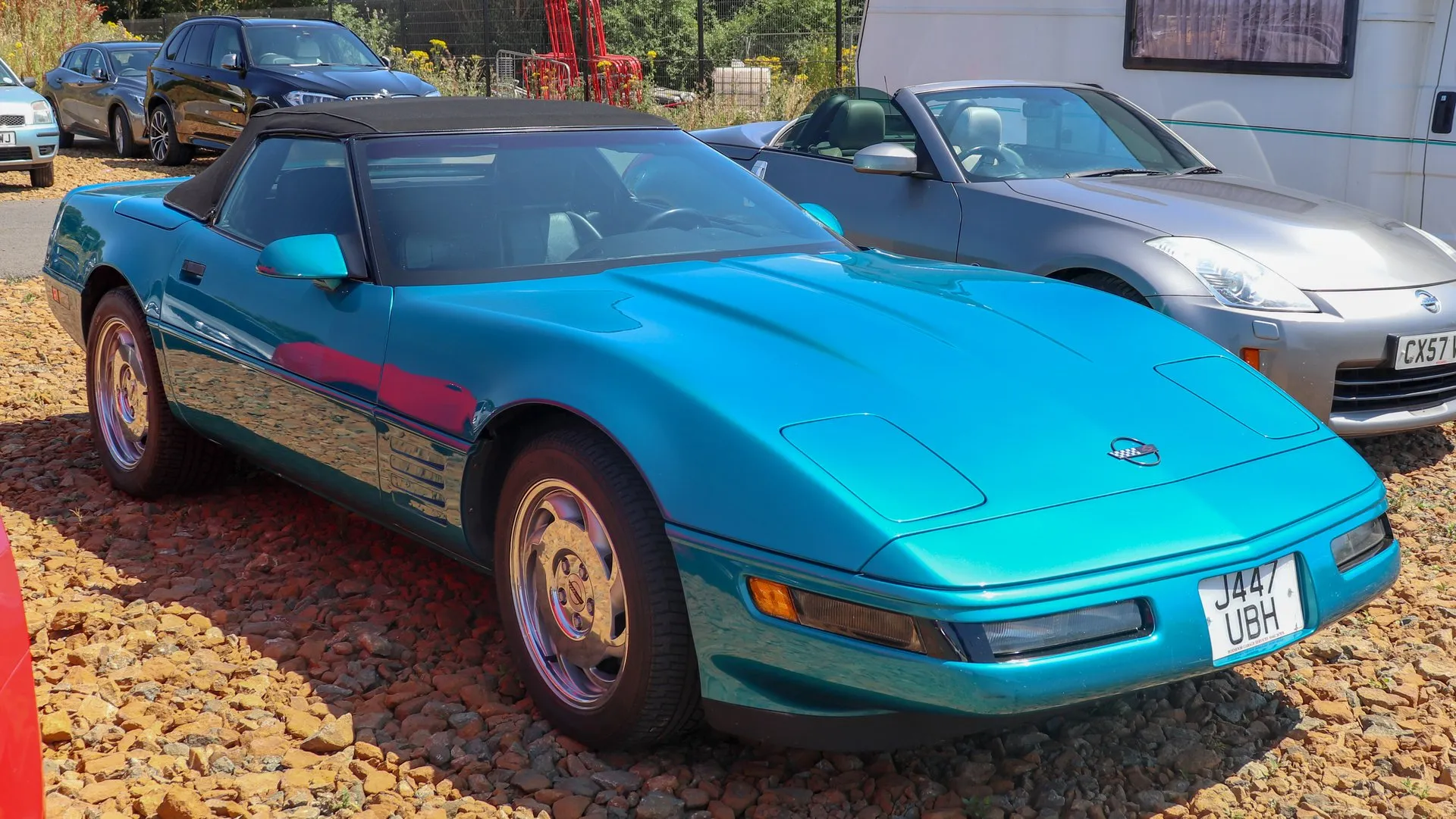 Bright Aqua Metallic Chevrolet Corvette C4 convertible with its black top up, parked on gravel at a car show and seen from the front right, with polished wheels and a British number plate.