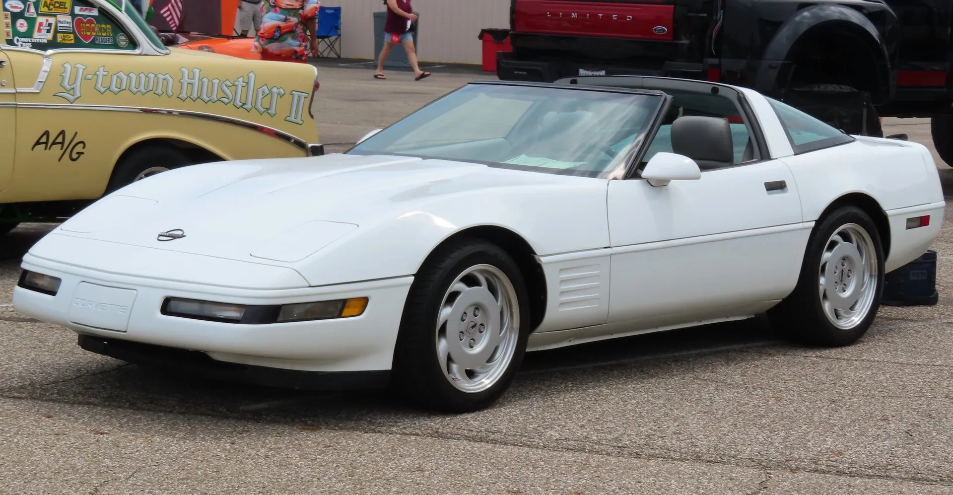 White Chevrolet Corvette C4 coupe with silver slotted wheels and a gray interior, parked on concrete at a car show and seen from the front left, beside an old drag racing car.