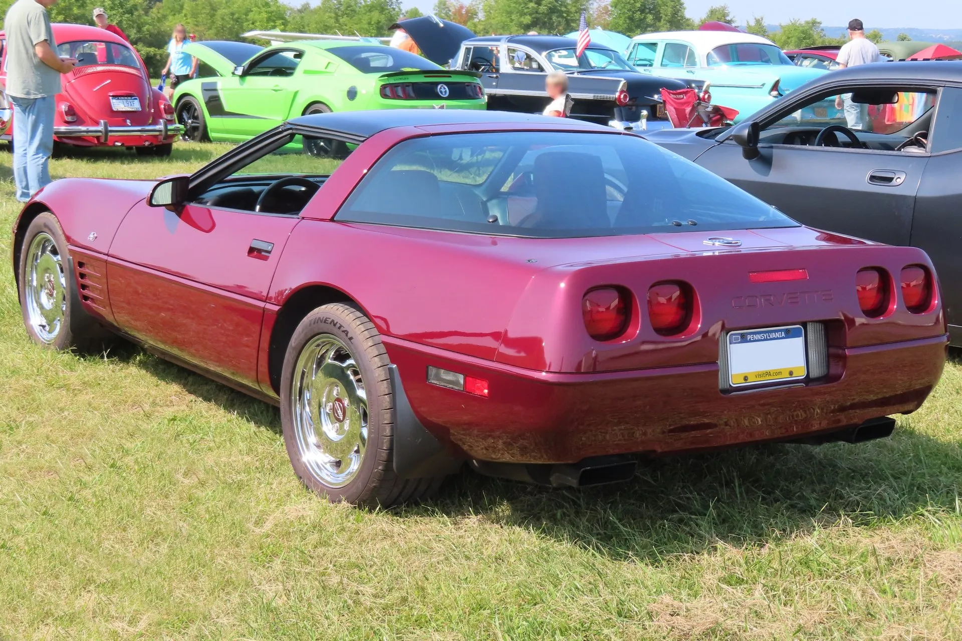 The Ruby Red coupe from the rear left at a car show, showing four round taillamps, Corvette lettering recessed into the rear fascia and two rectangular exhaust outlets.