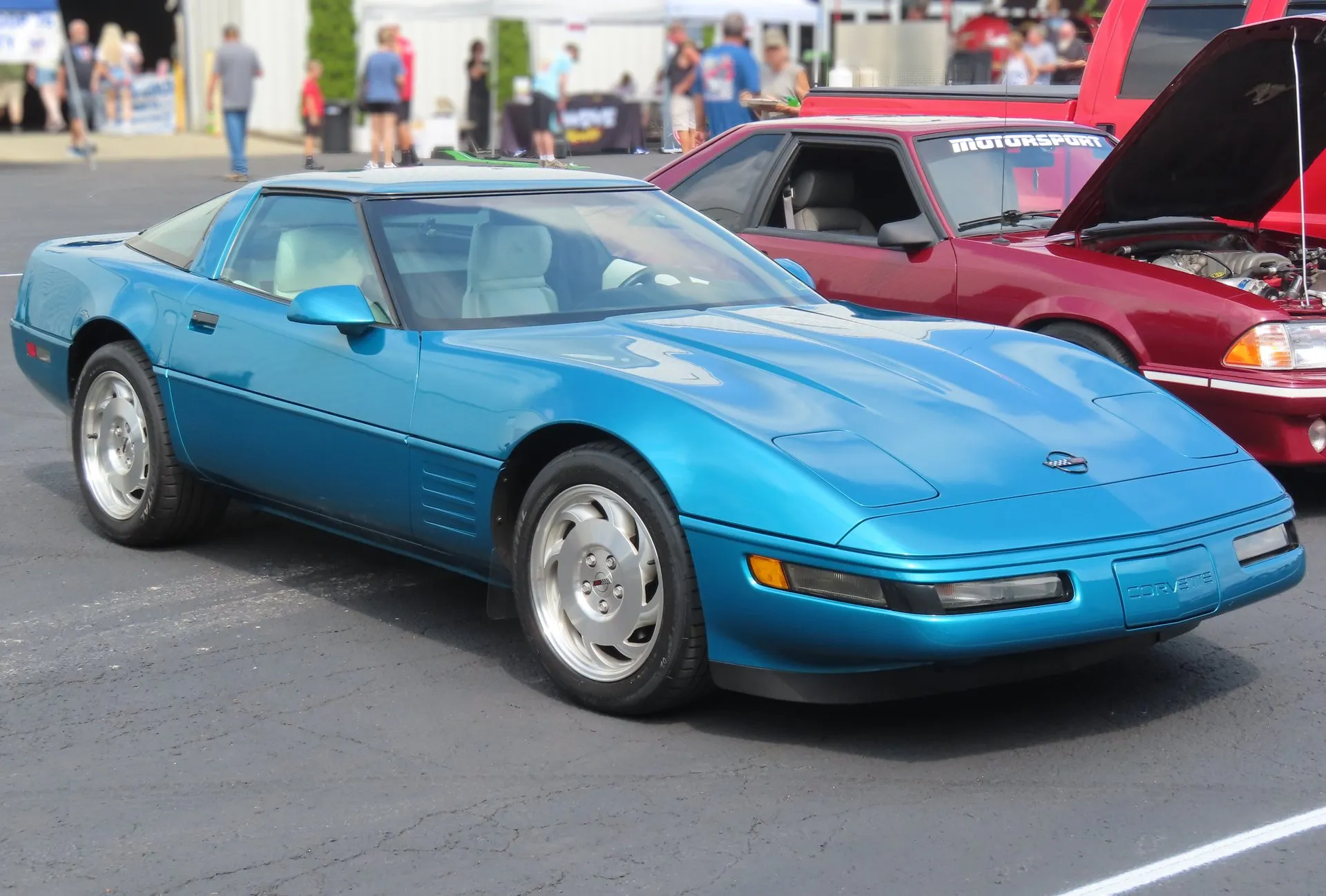 Bright Aqua Metallic C4 Corvette coupe with a light interior and silver slotted wheels, parked on asphalt at a car show, seen from the front right.