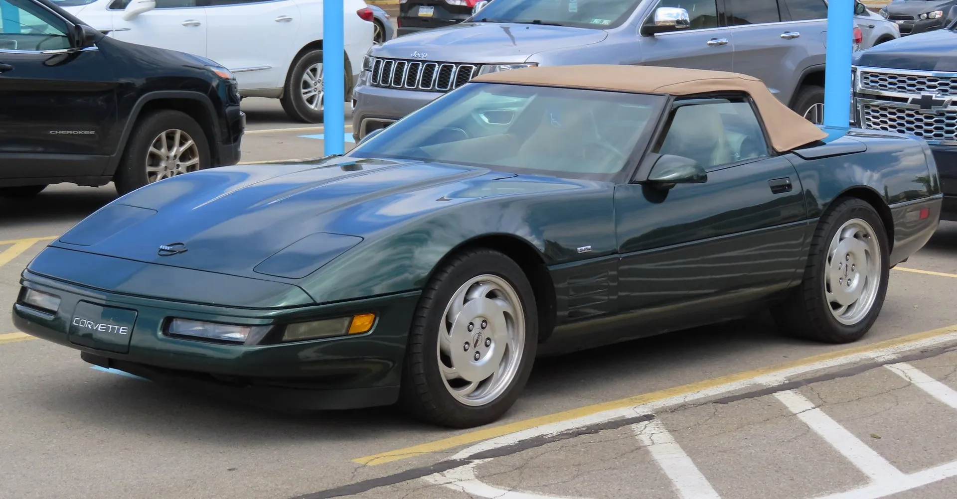 Dark green C4 Corvette convertible with a tan top up, in a parking lot among newer cars, seen from the front left, with silver wheels and straight strakes behind the front wheel.