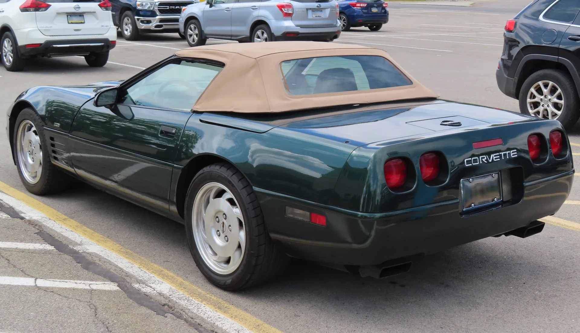 The same dark green C4 Corvette convertible from the rear left with its tan top up, showing the top's rear window, four round red taillamps and Corvette lettering on the rear fascia.